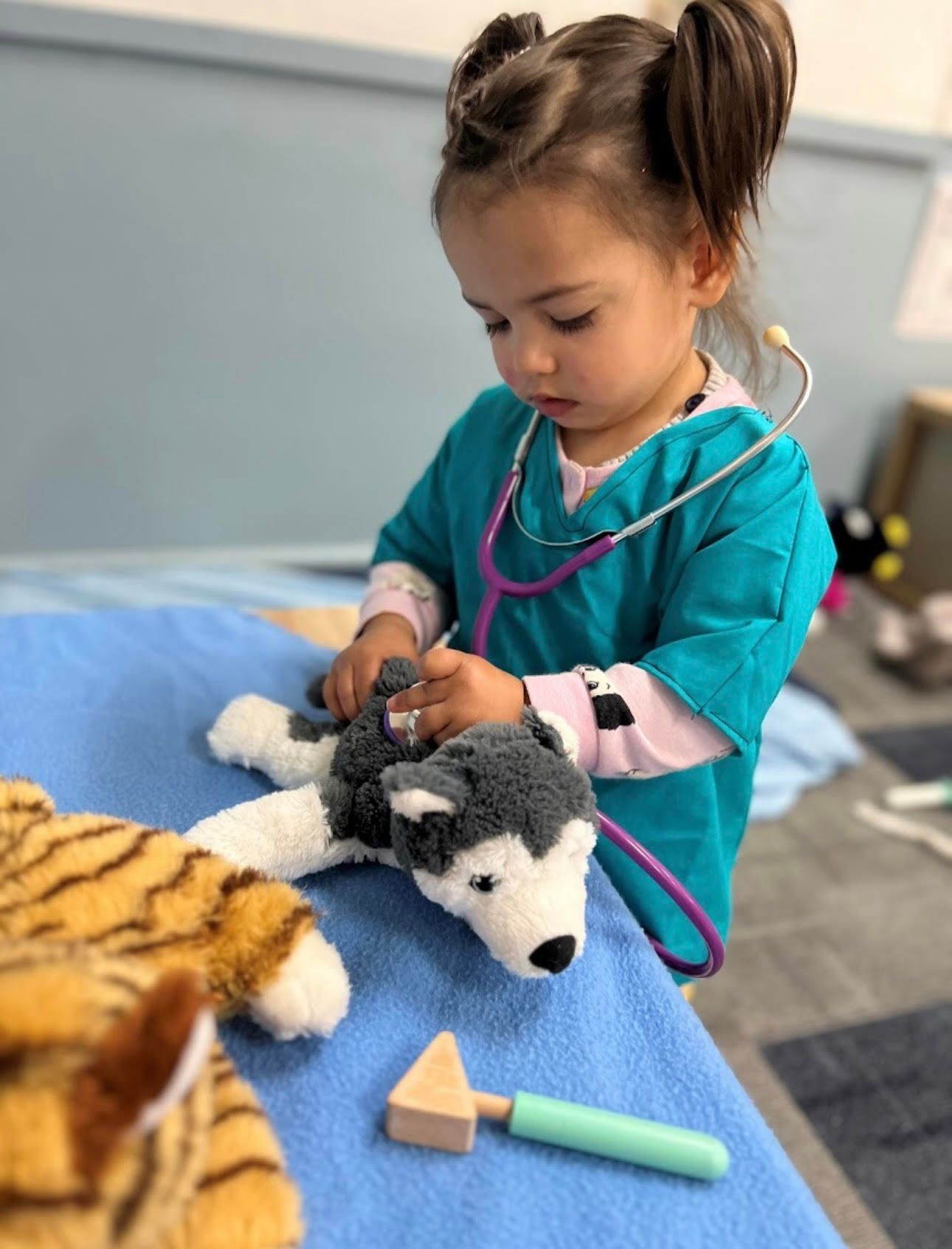A young child in a blue medical uniform examines a stuffed animal with a stethoscope on a table.