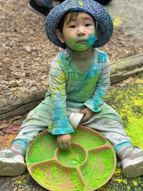 Child covered in colorful powder sits with a tray, smiling. Wearing a hat and light-colored clothes. Outdoors.