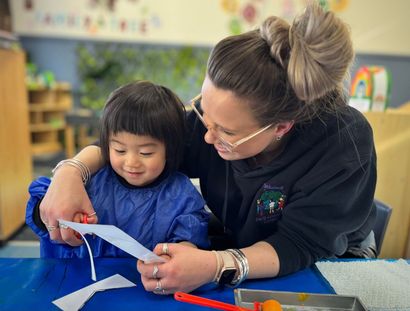 Woman helps a child cut paper with scissors at a blue table in a classroom.