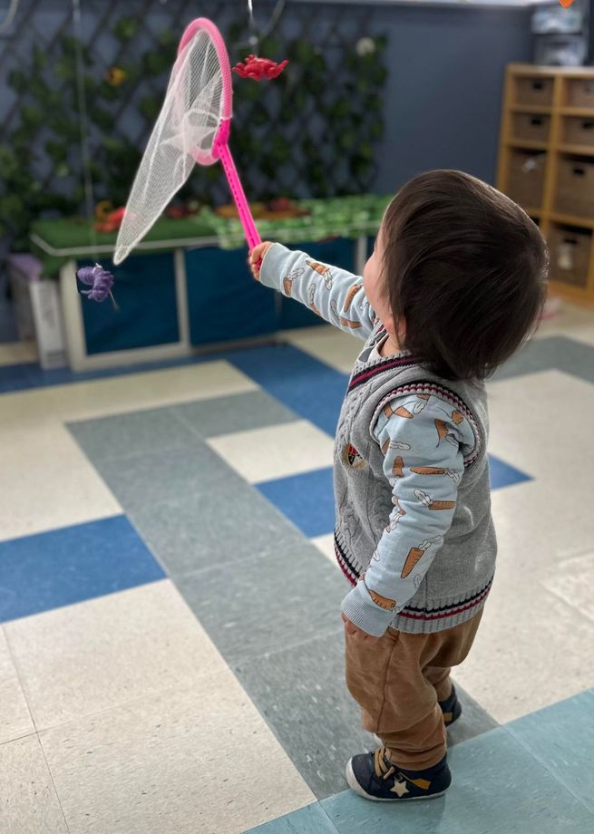 Child in a vest holds up a pink butterfly net inside a room with a decorative butterfly.