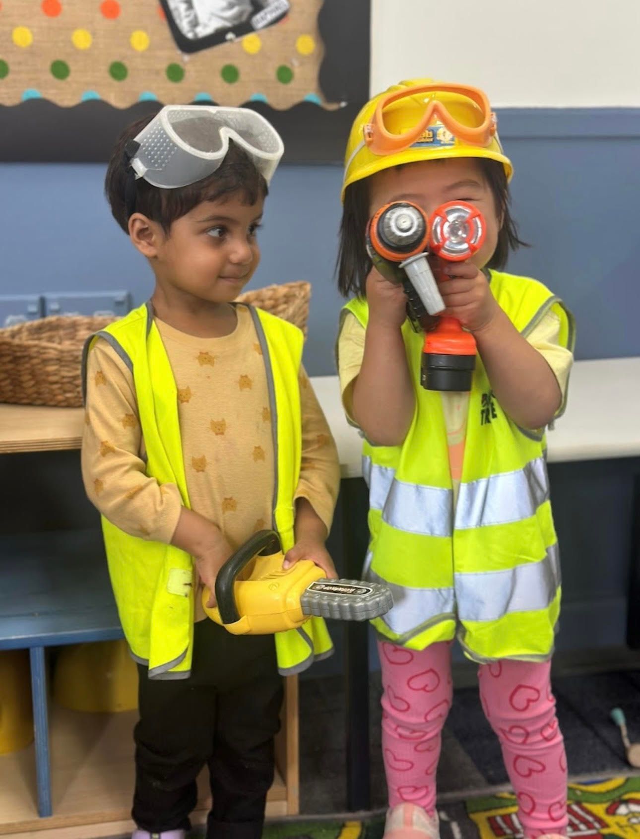 Two children in construction gear: one with a toy saw, the other with a toy drill, both smiling.