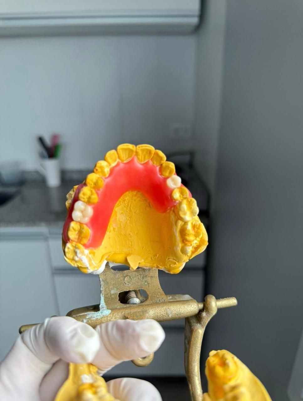 Mano sosteniendo un modelo dental de dientes con encías de cera y cera rosa, montado en un laboratorio dental.