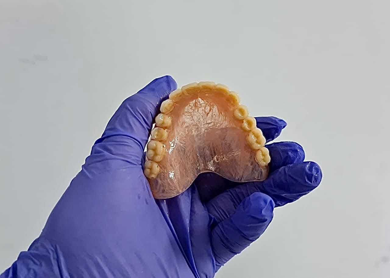 Mano enguantada sosteniendo una dentadura postiza superior, con los dientes hacia el espectador. El fondo es blanco liso.