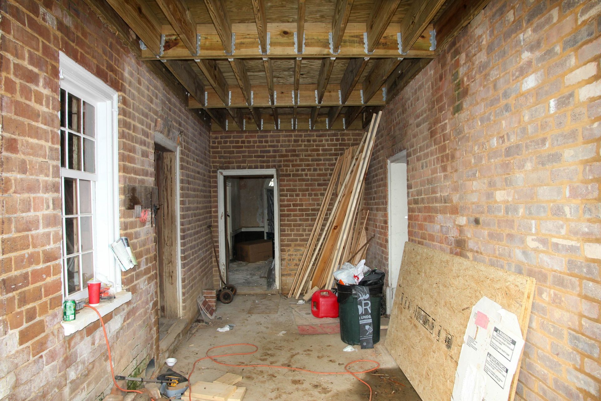 Unfinished brick corridor with exposed ceiling beams, construction materials, and a doorway at the far end.