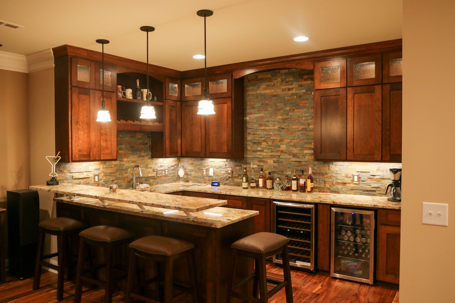 A home bar with wooden cabinets, stone backsplash, granite countertops, bar stools, and two wine fridges.