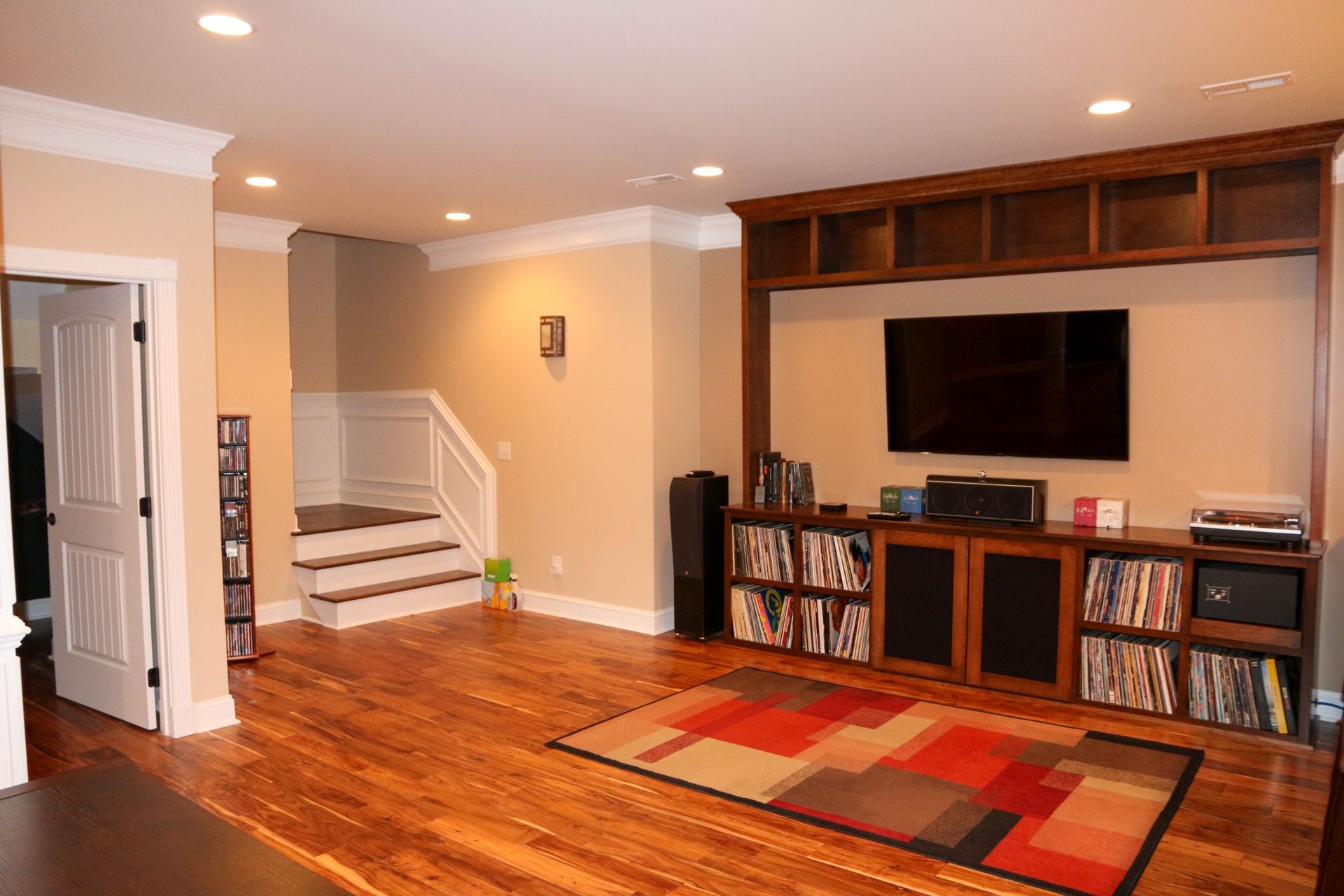 Spacious living room with hardwood floors, wall-mounted TV, shelves, staircase, and red patterned rug.