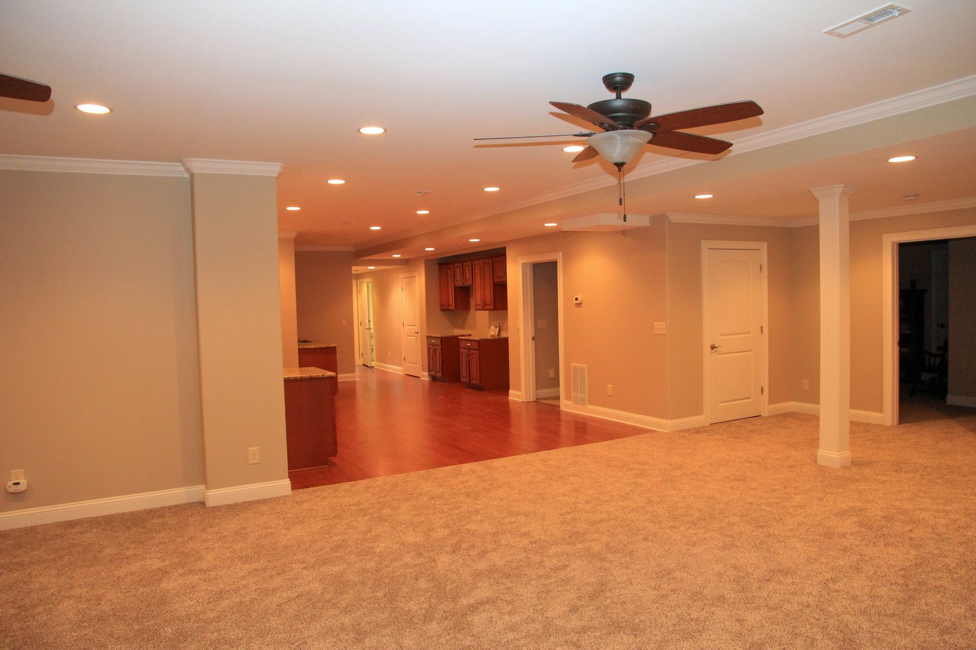 Empty beige living room with carpet, recessed lights, and a ceiling fan over wood flooring.