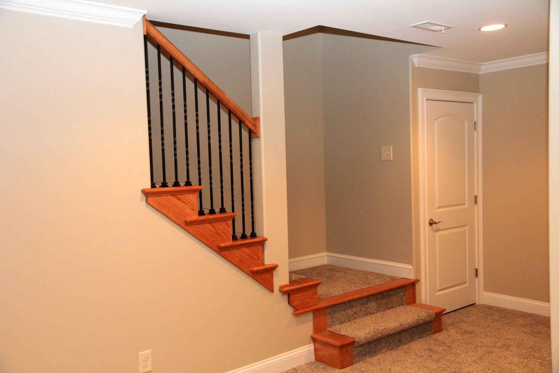 Indoor staircase with dark railings, red-brown steps, beige walls, and a closed white door beside carpeted flooring