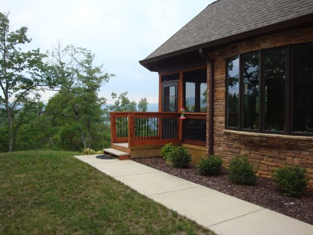 Stone house exterior with wooden porch, large windows, and a concrete walkway beside a grassy lawn