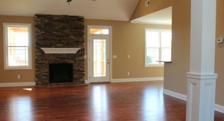 Empty living room with hardwood floors, stone fireplace, beige walls, and large windows with natural light