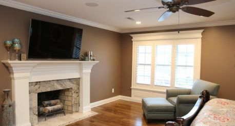 Living room with white fireplace, wall-mounted TV, hardwood floors, and gray chair by a bright window.