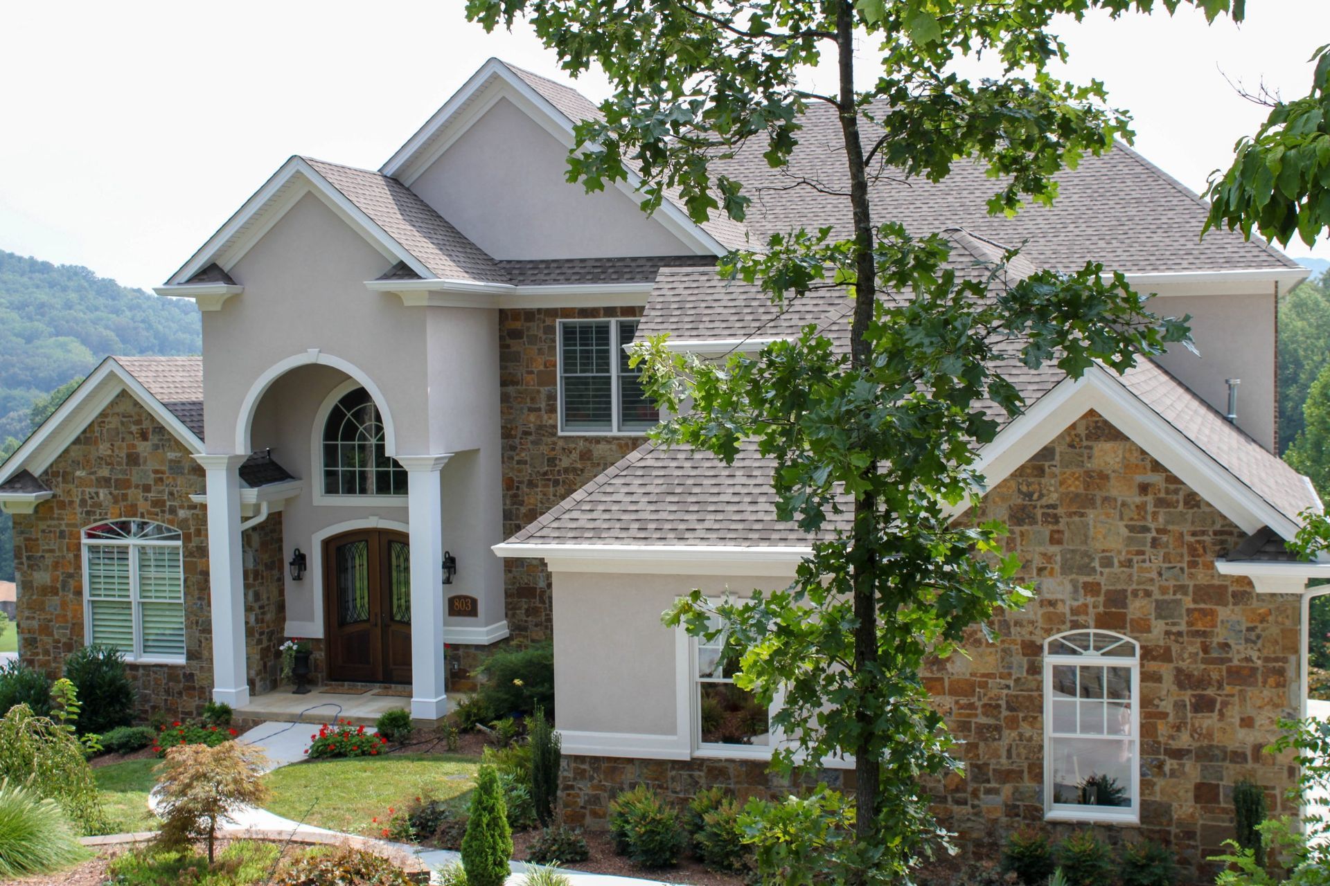 Large suburban stone house with arched entry, gray roof, and landscaped front yard