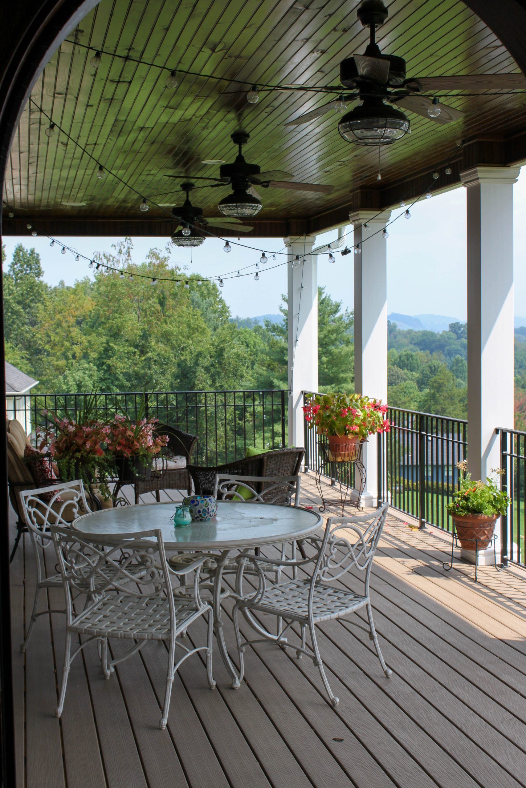 Covered patio with white metal table and chairs, ceiling fans, potted flowers, and a green mountain view.
