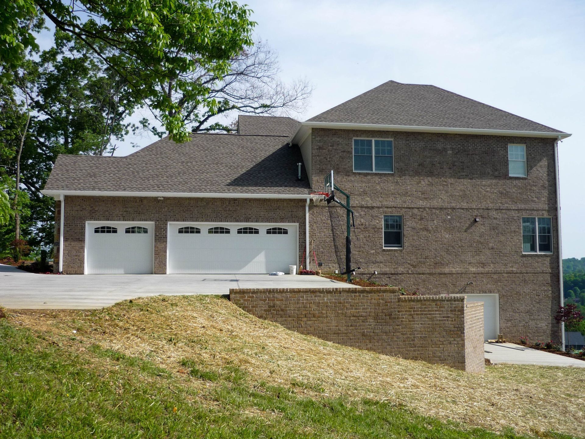 Brick suburban house with three-car garage, stone retaining wall, and sloped lawn under a tree
