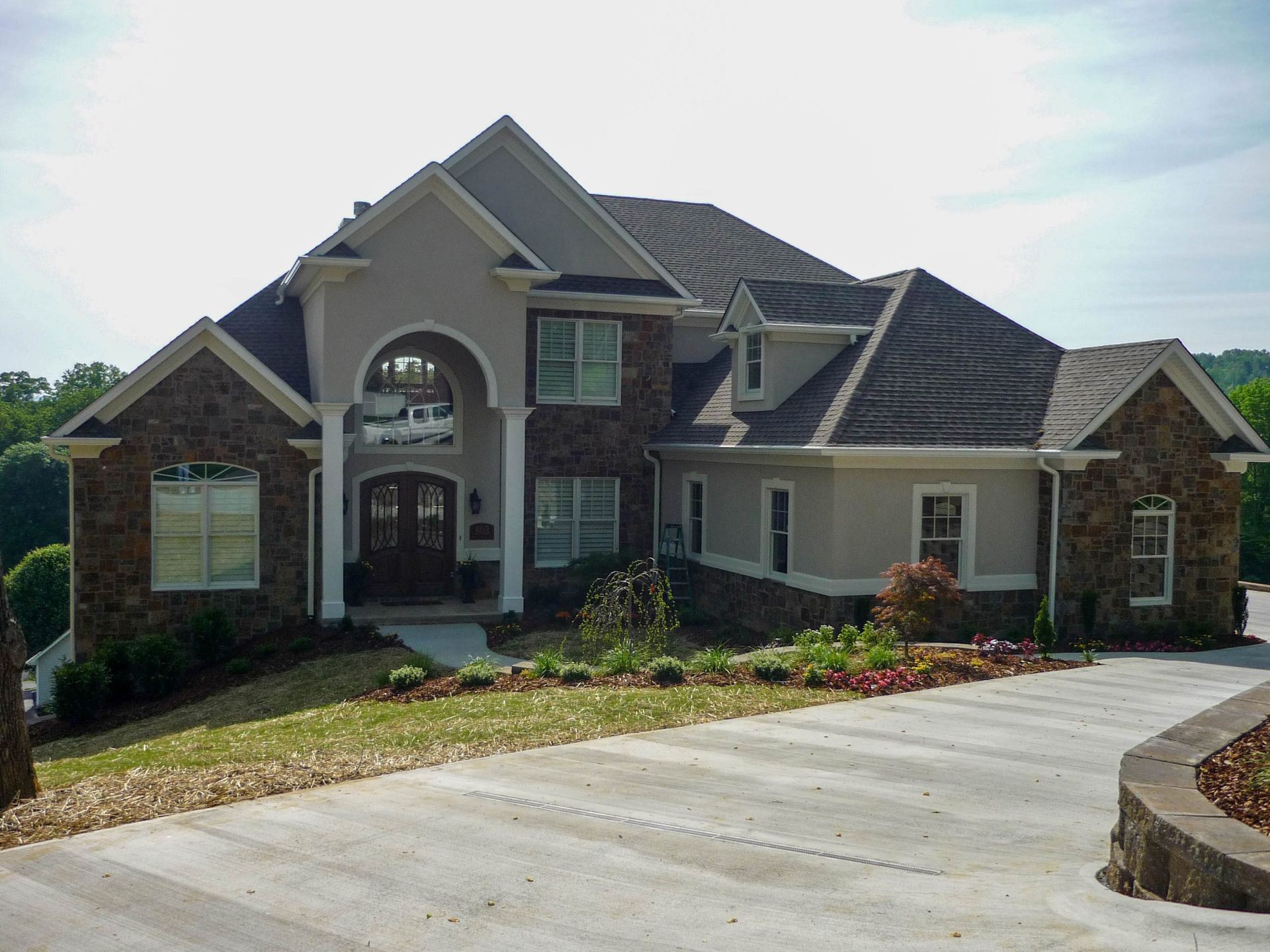 Large brick suburban house with arched entry, gray roof, and wide driveway on a landscaped hillside