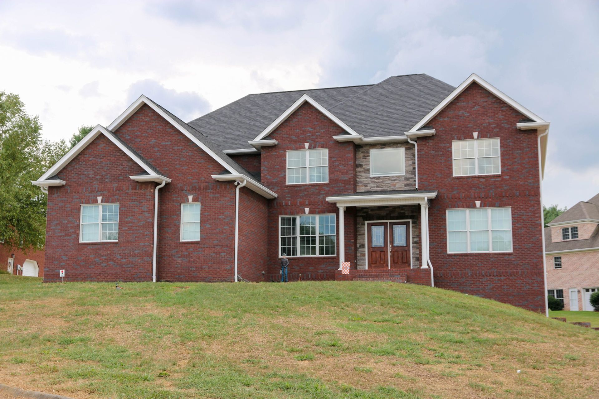 Large red-brick two-story house with white trim on a grassy hill under a cloudy sky