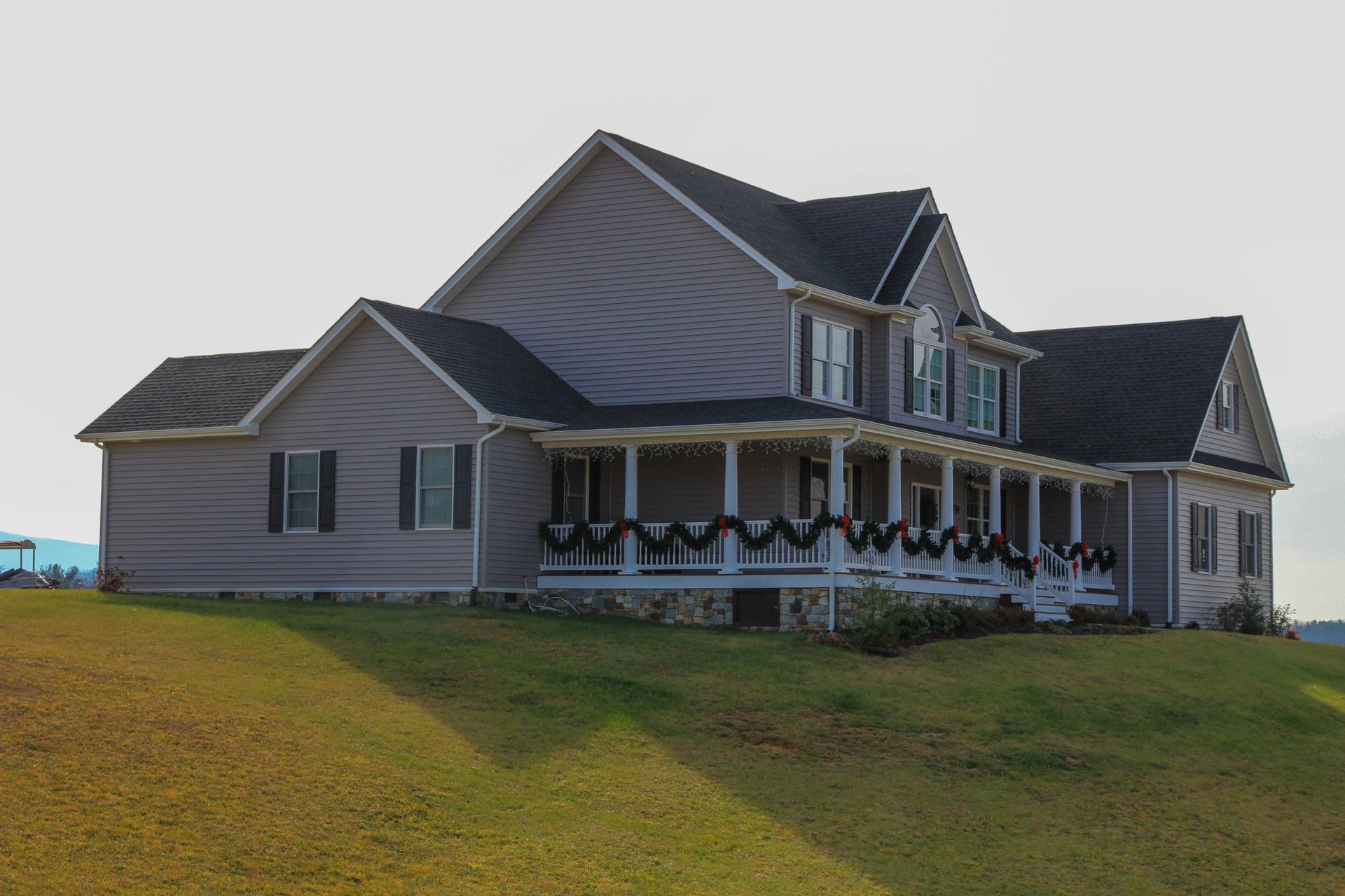 Large gray two-story house with a wraparound porch on a grassy hill under a cloudy sky