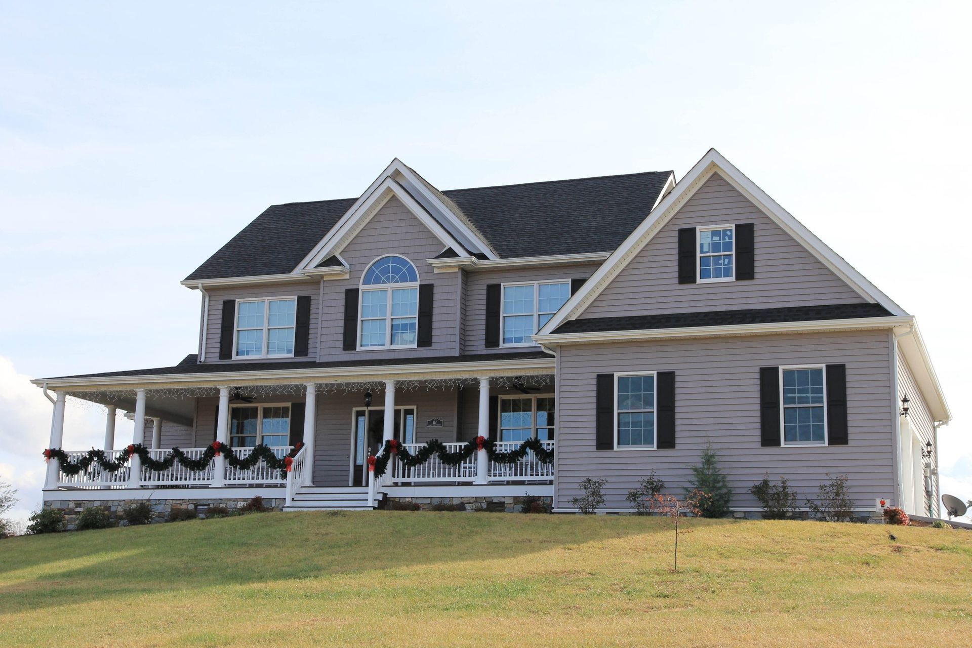 Two-story beige house with a front porch, dark roof, and holiday garland on a grassy hill