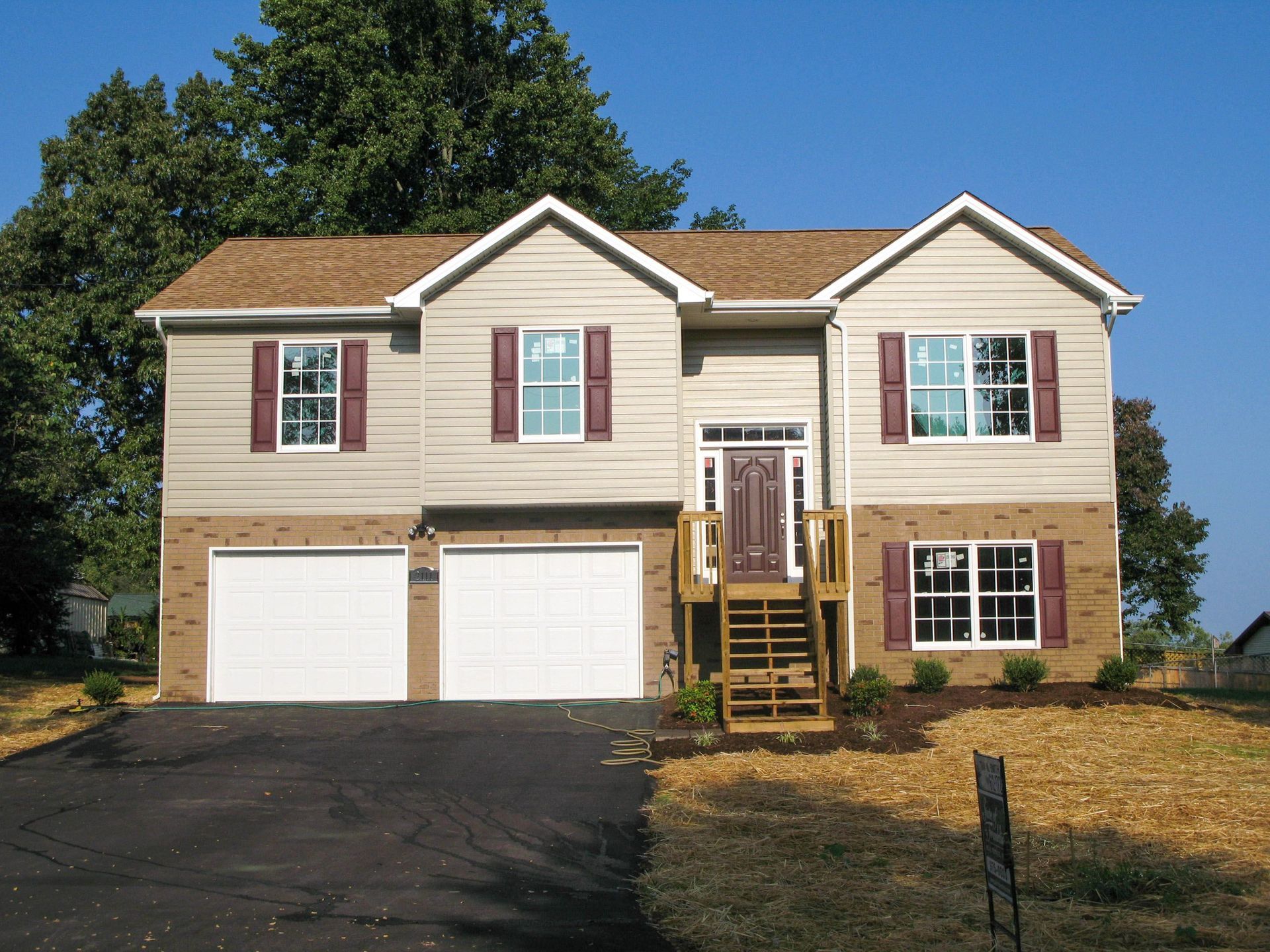 Suburban two-story beige house with white garage doors, front porch, and a driveway under a clear blue sky