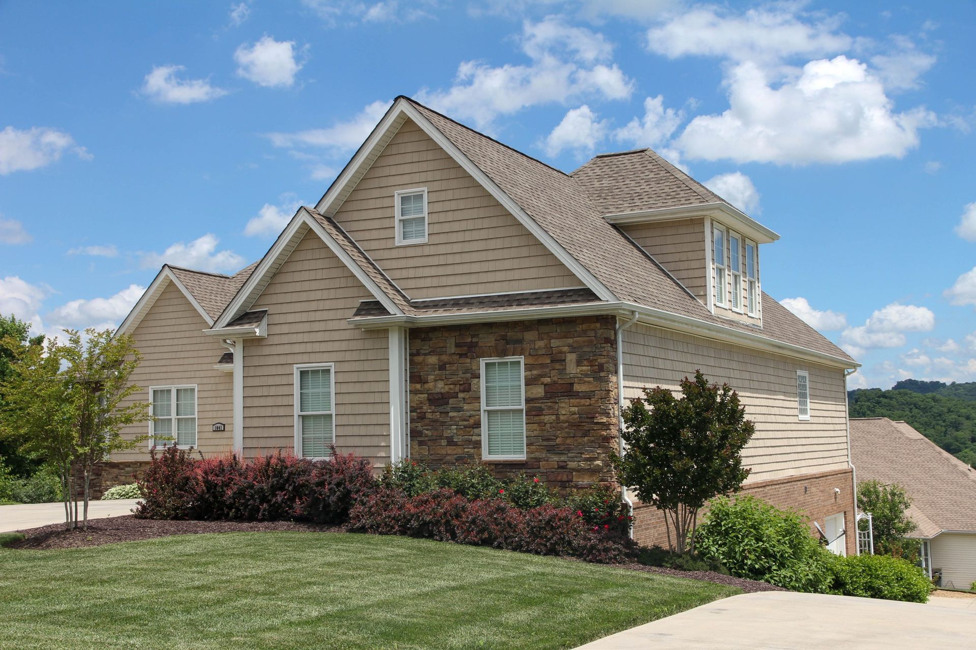 Suburban beige brick house with shingled roof, landscaped yard, and blue sky.