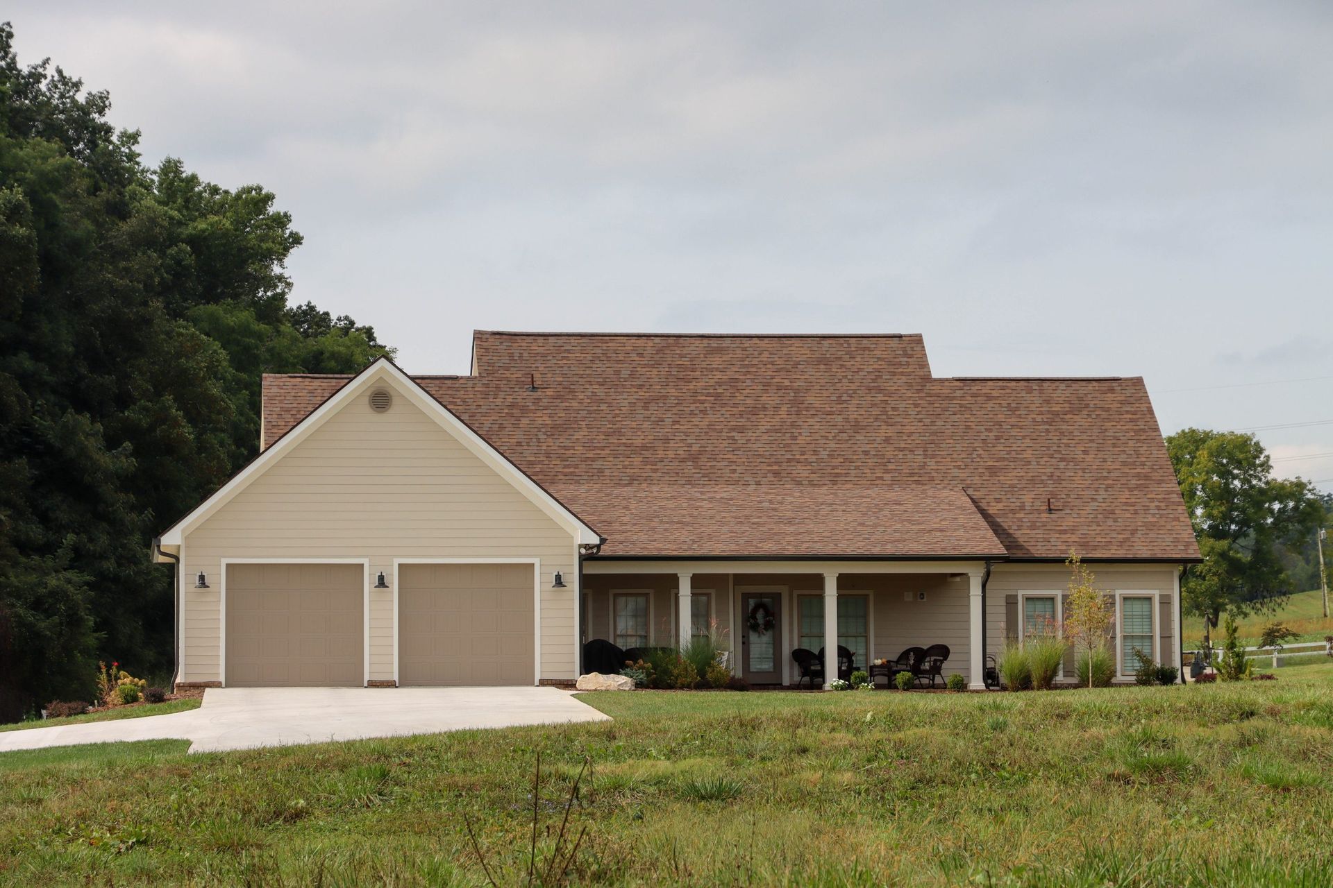 Single-story beige house with a brown roof, front porch, and attached two-car garage on a grassy lot