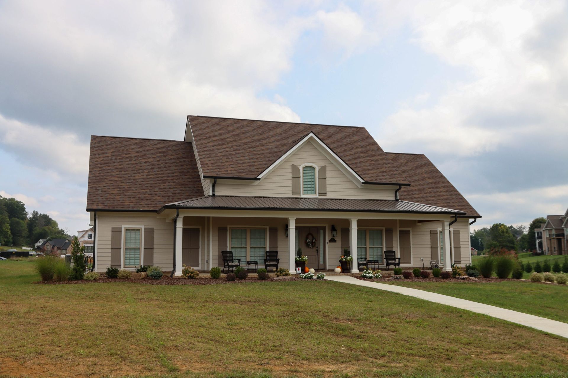 A beige suburban house with a front porch, brown roof, and a grassy yard under a cloudy sky.