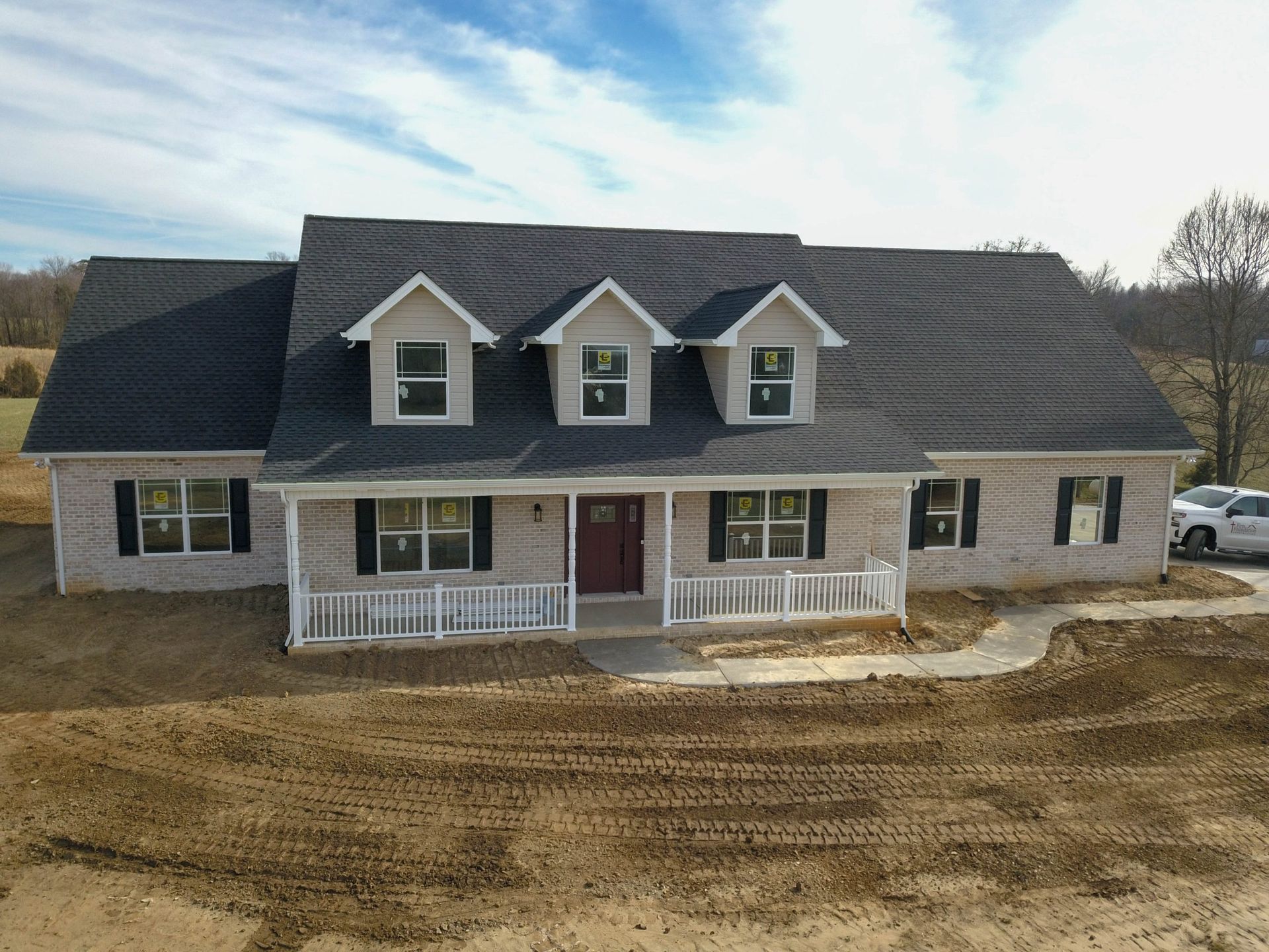Two-story house with gray roof, white porch railings, and dormer windows on a bare dirt lot.