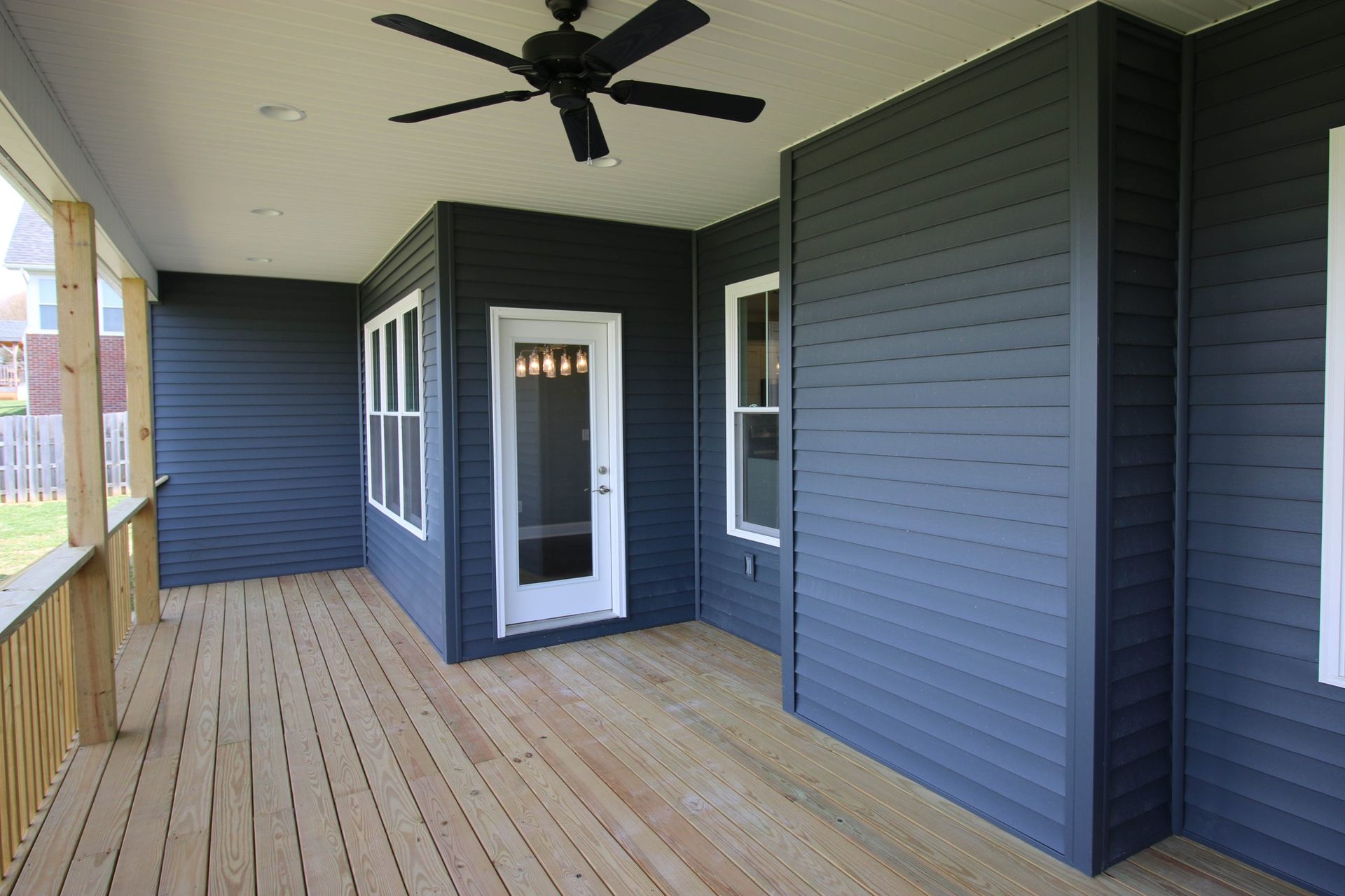 Covered wooden porch with dark blue siding, white door, and ceiling fan.
