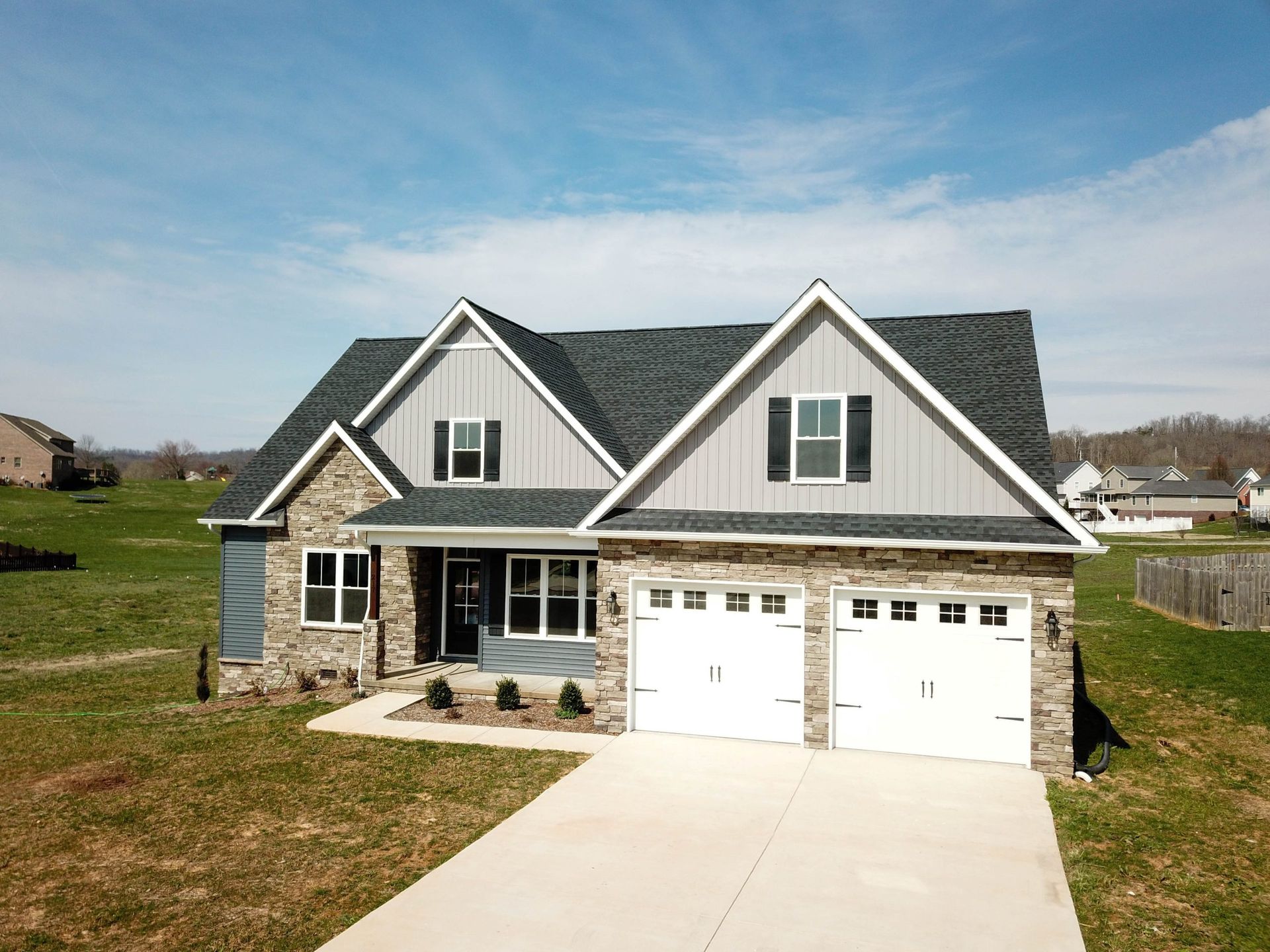 Two-story suburban house with gray stone facade, white garage doors, and a wide driveway under a blue sky