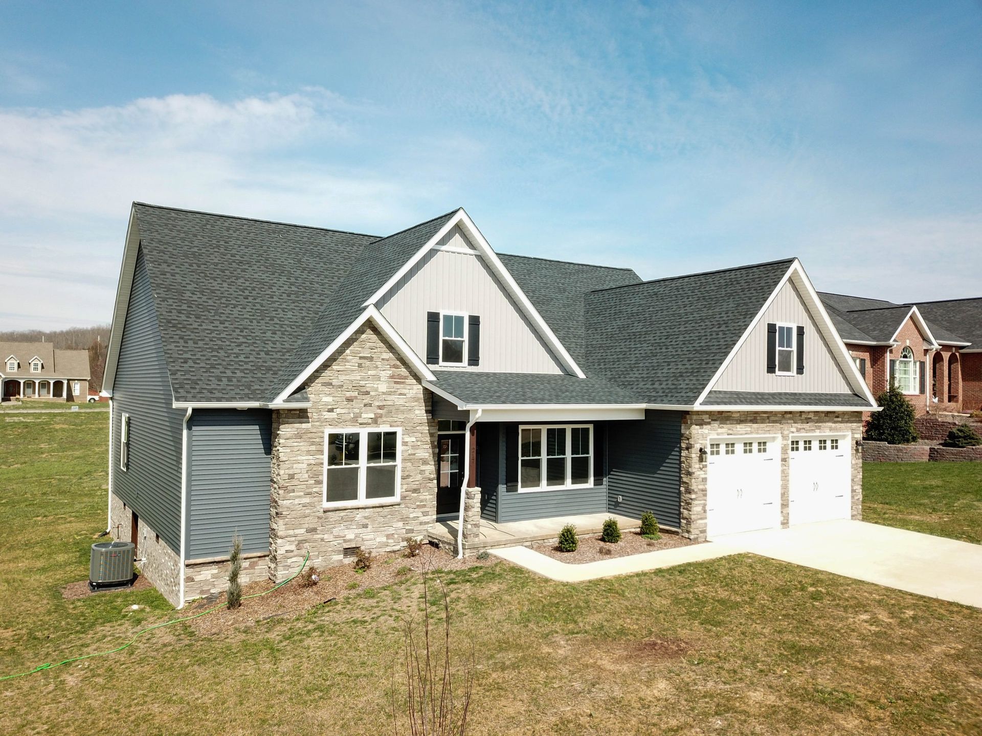 Suburban stone house with gray roof, white garage doors, and a front lawn under a blue sky