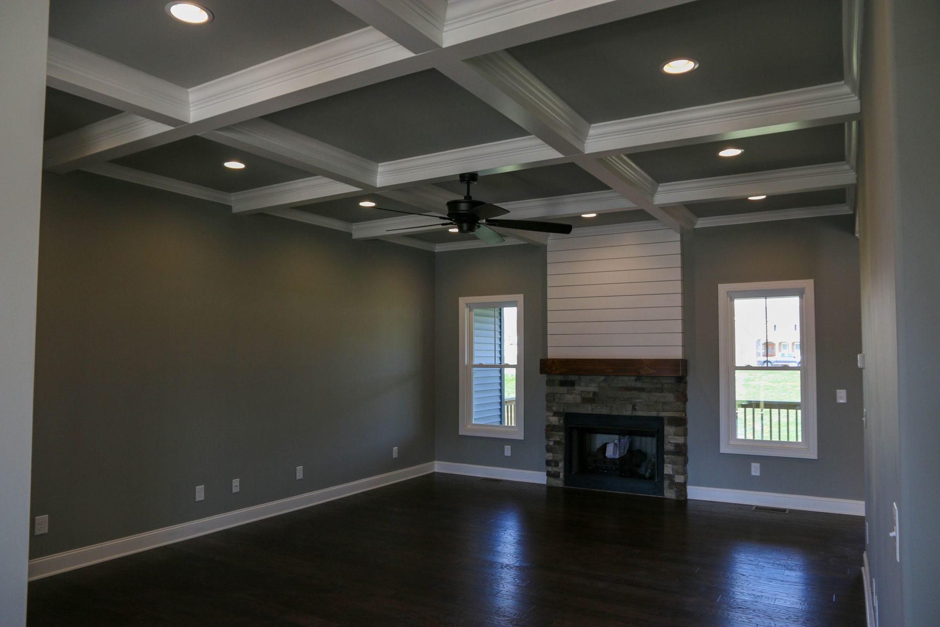 Empty living room with dark wood floors, coffered ceiling, fireplace, and two windows.