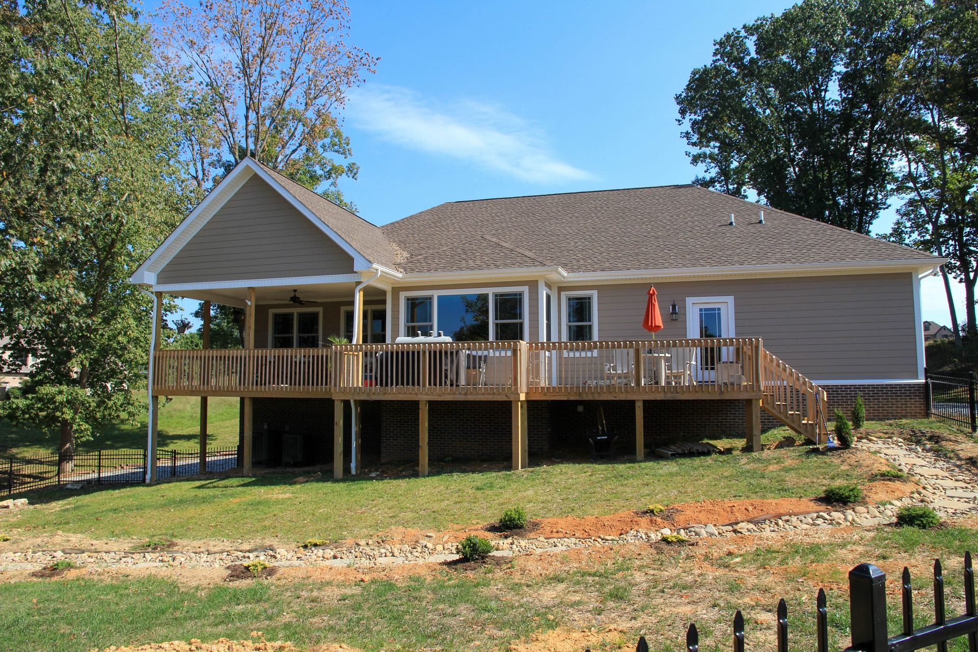 Single-story tan house with a large wooden deck and stairs in a grassy yard, surrounded by trees.