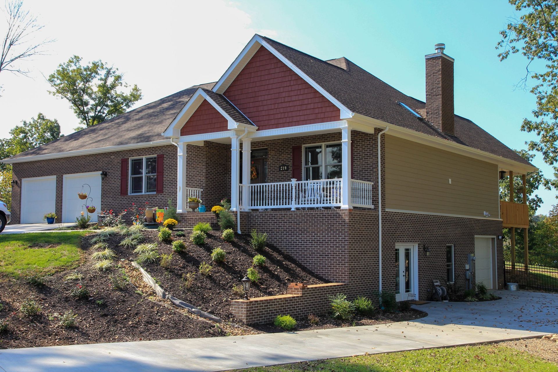 House with red siding, tan walls, white porch railing, and a landscaped sloped yard in sunlight