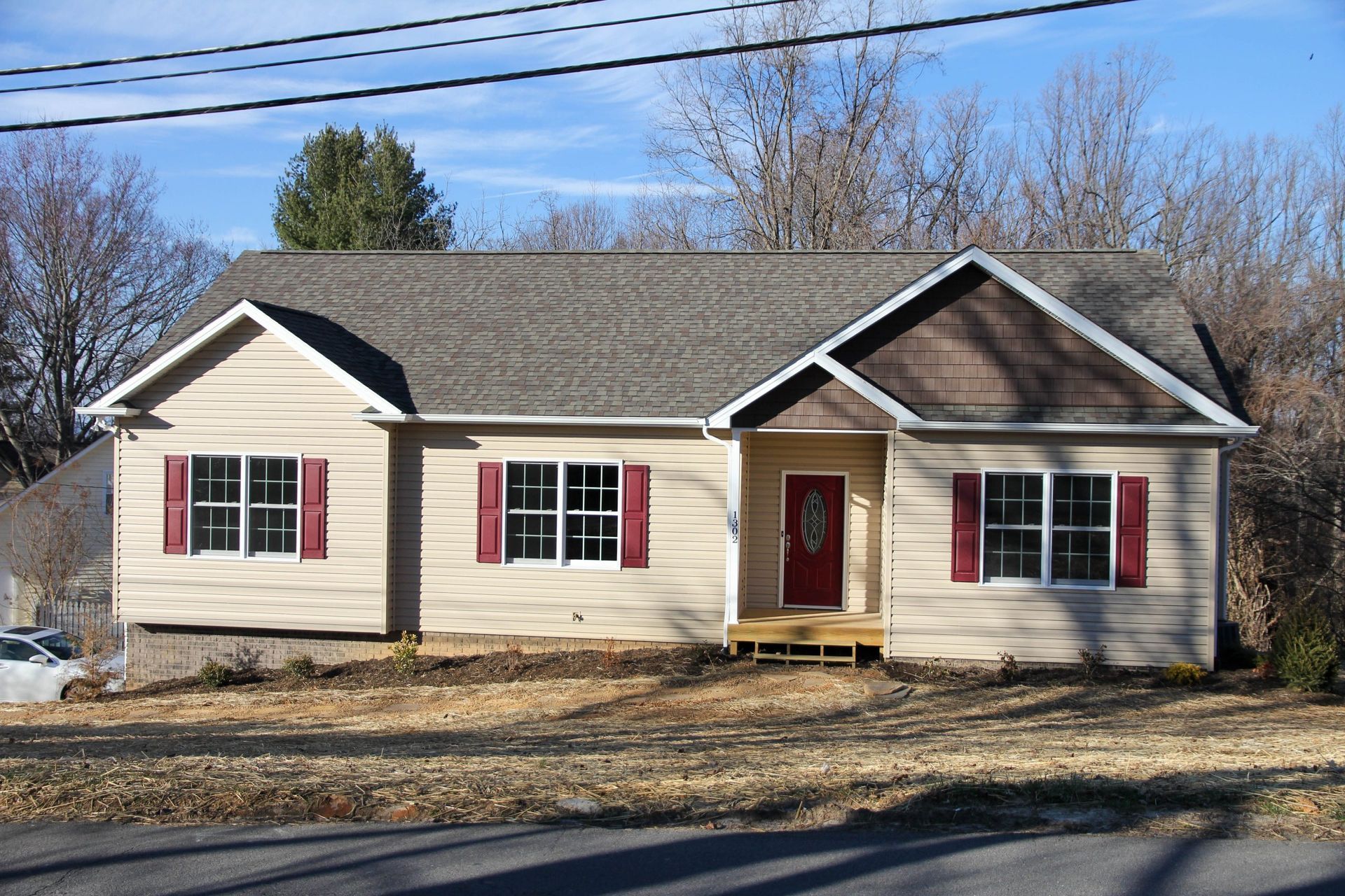 Single-story beige house with red shutters and a red front door, set beside a road under bare trees.