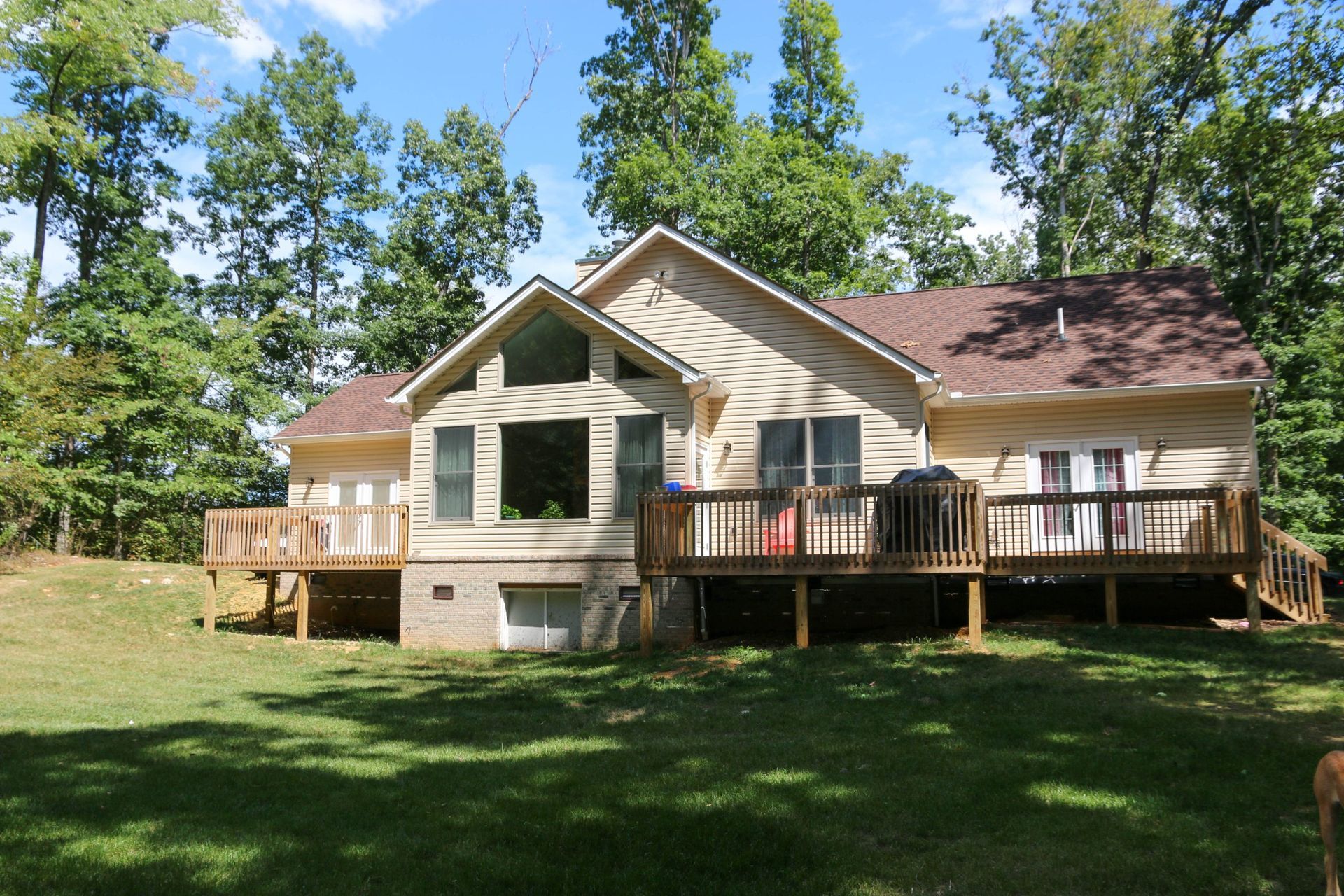 Single-story beige house with wooden decks in a grassy, wooded yard under a blue sky