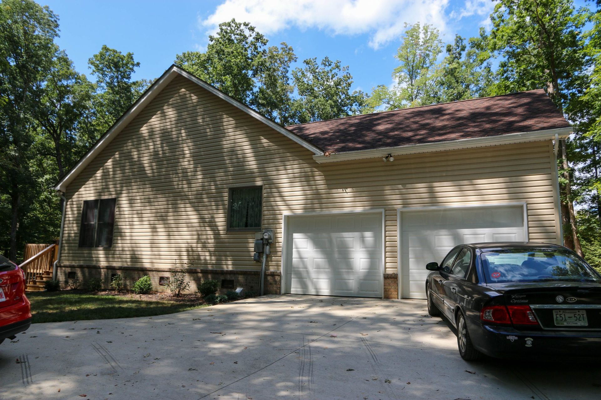 Suburban house with beige siding, two white garage doors, driveway, and parked cars under trees