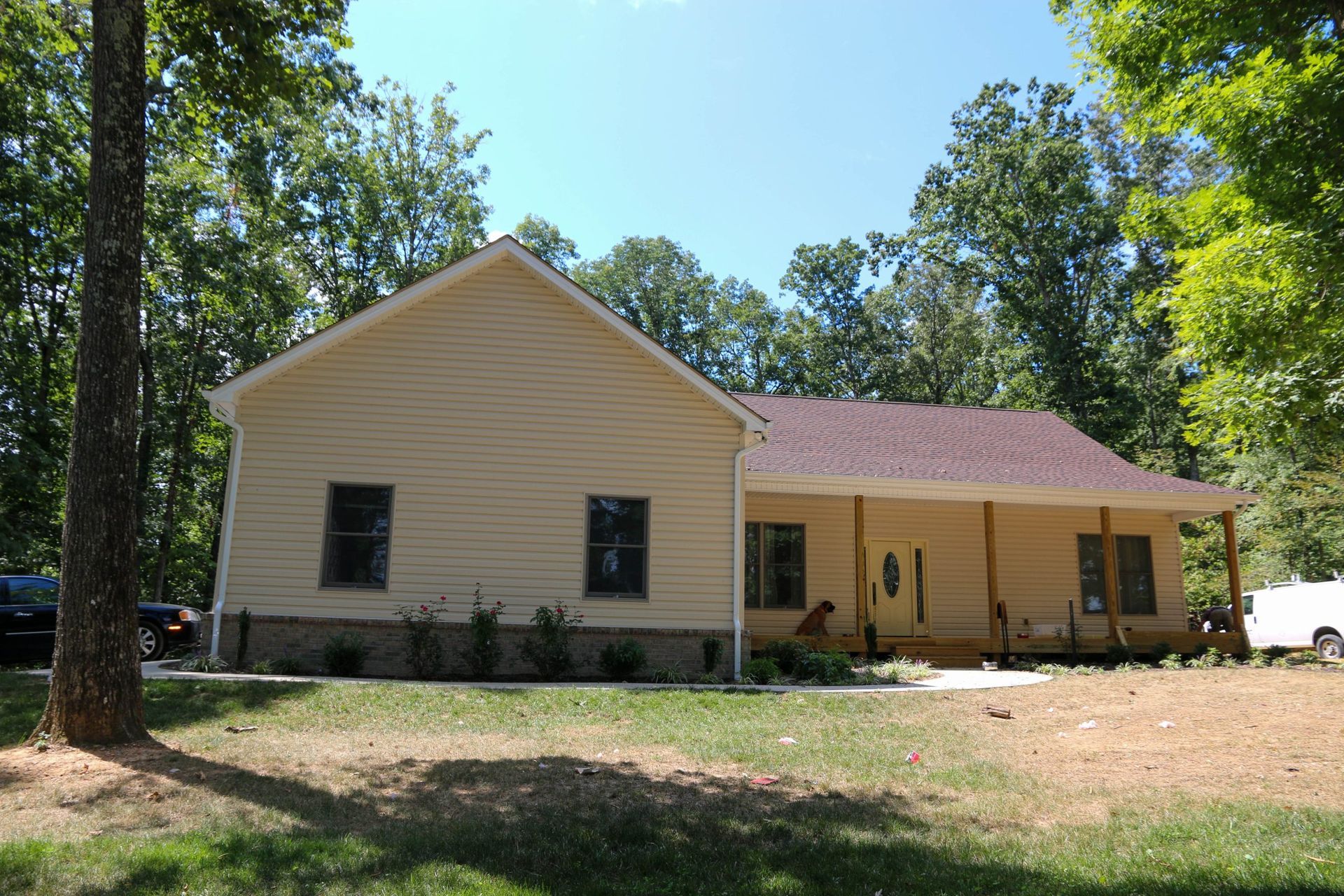 Tan house with green shutters and a porch in a wooded yard on a sunny day