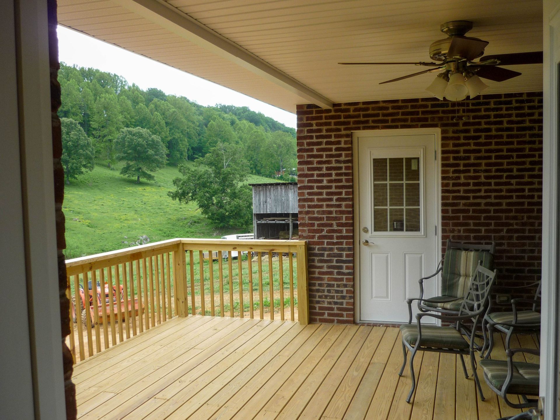 Covered wooden deck with railing, white door, wicker chairs, and green hillside beyond.
