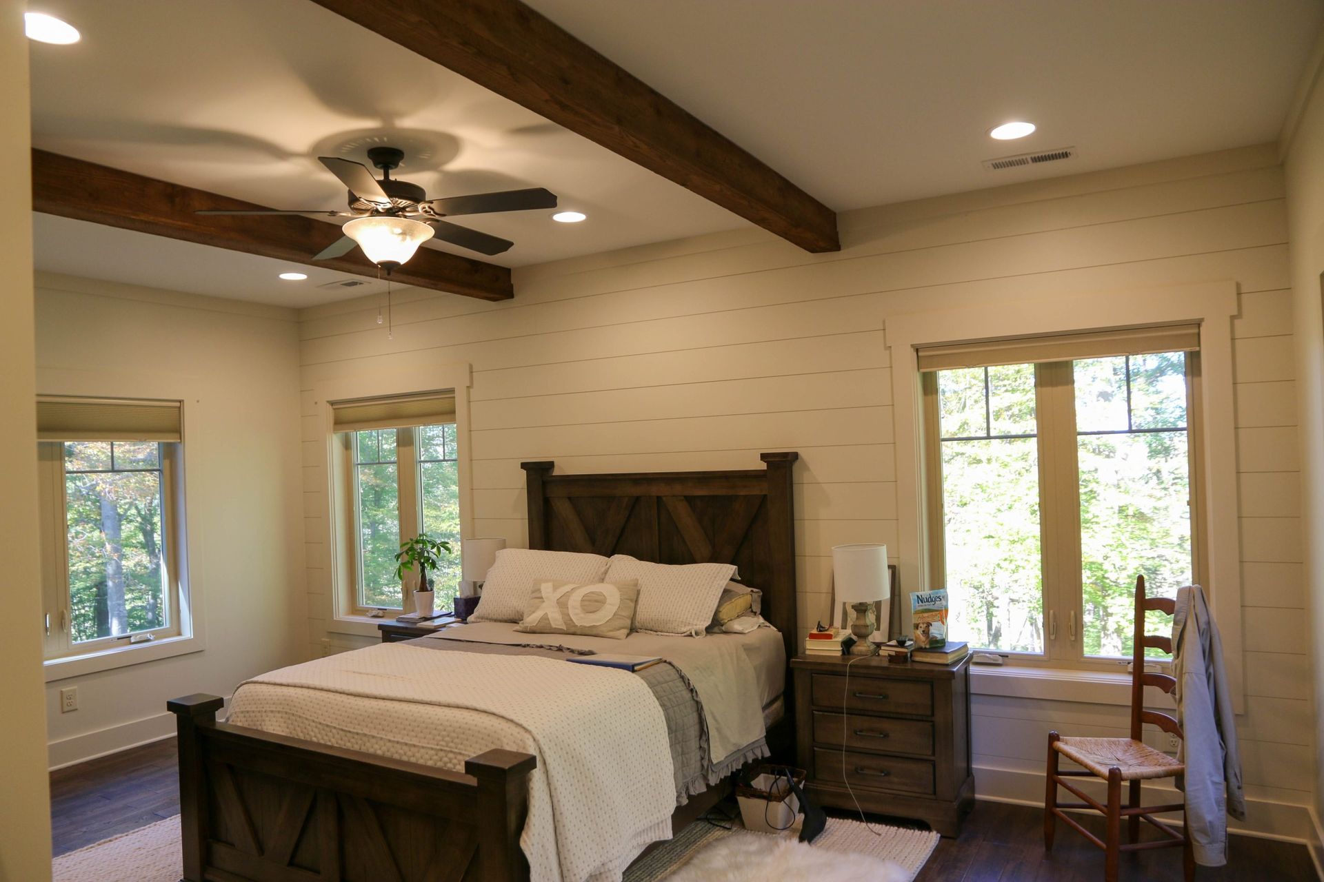 Bright bedroom with exposed beams, a dark wooden bed, ceiling fan, and large windows with natural light.