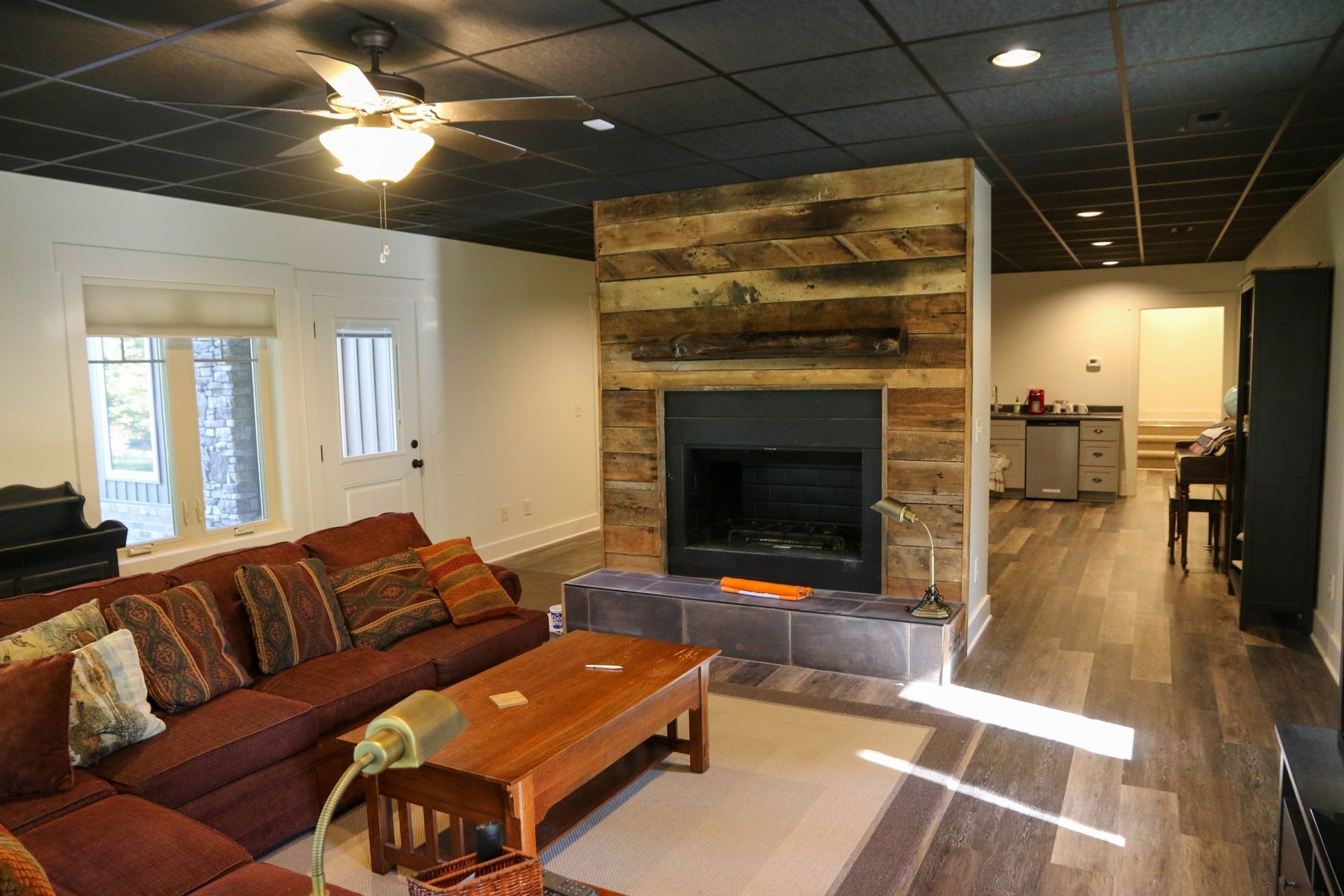 Living room with brown sectional sofa, stone fireplace, wood coffee table, and open view to kitchen