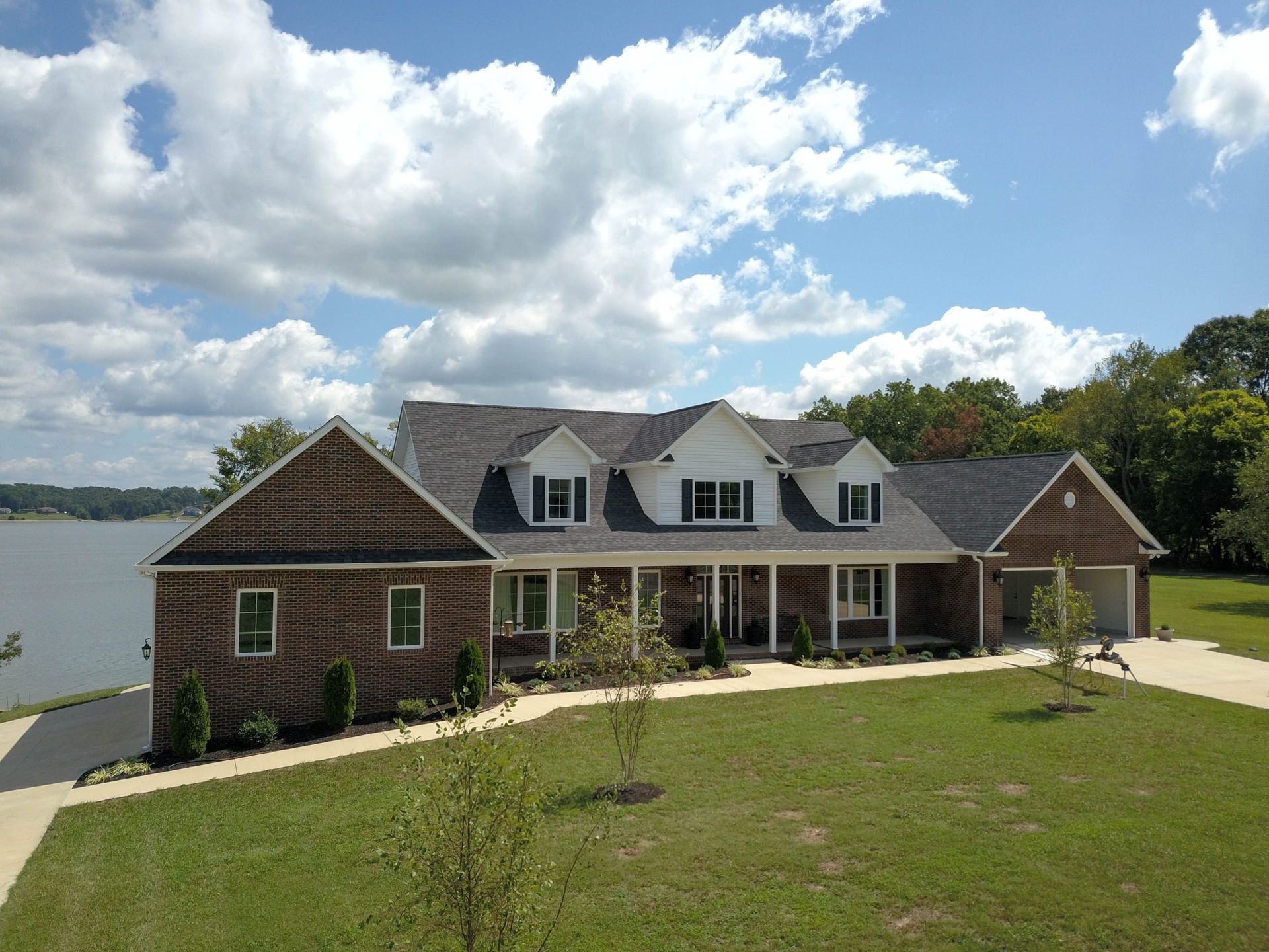 Lakefront house with gray roof, white porch, and green lawn under a cloudy blue sky