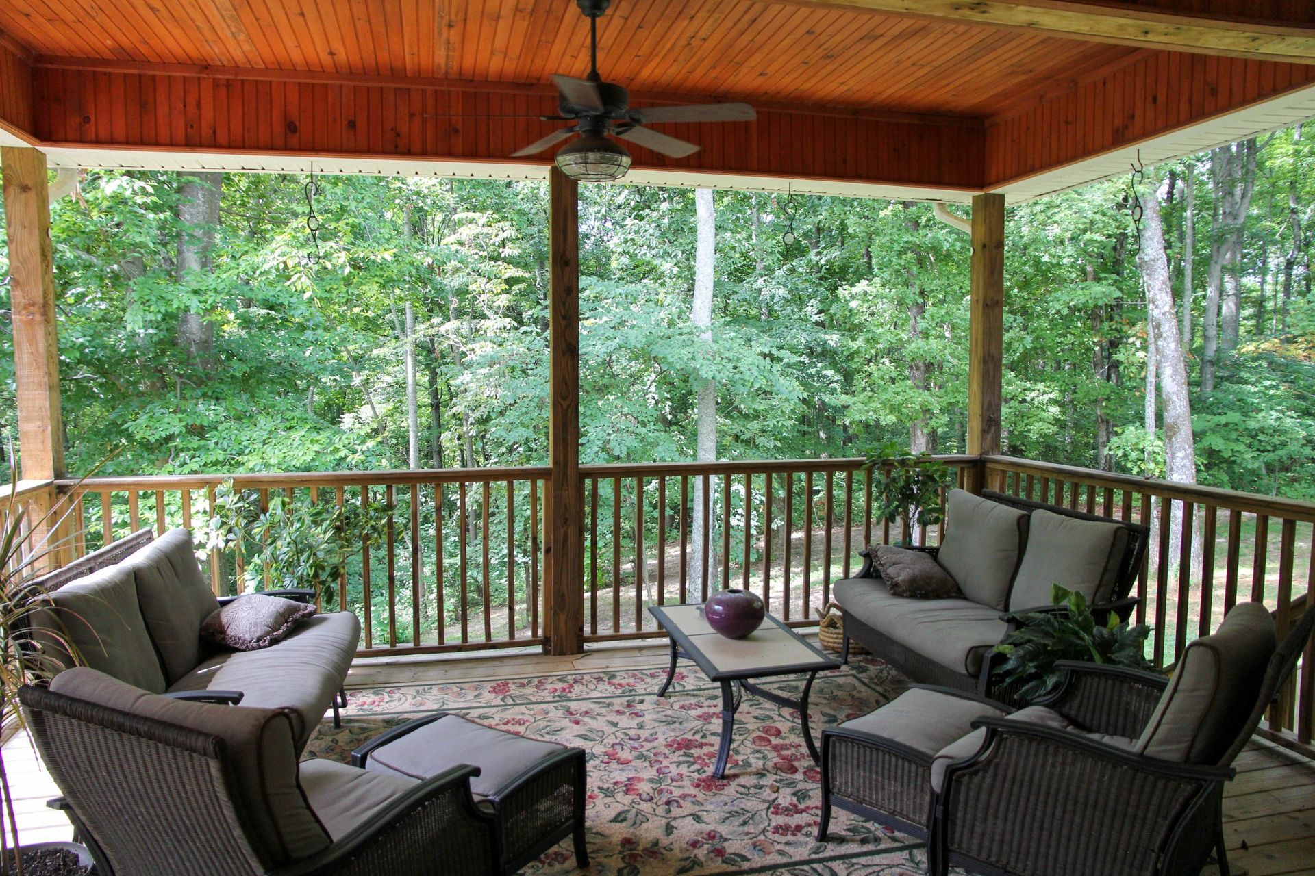 Covered porch with wicker seating, patterned rug, and wooded forest view beyond the railing.