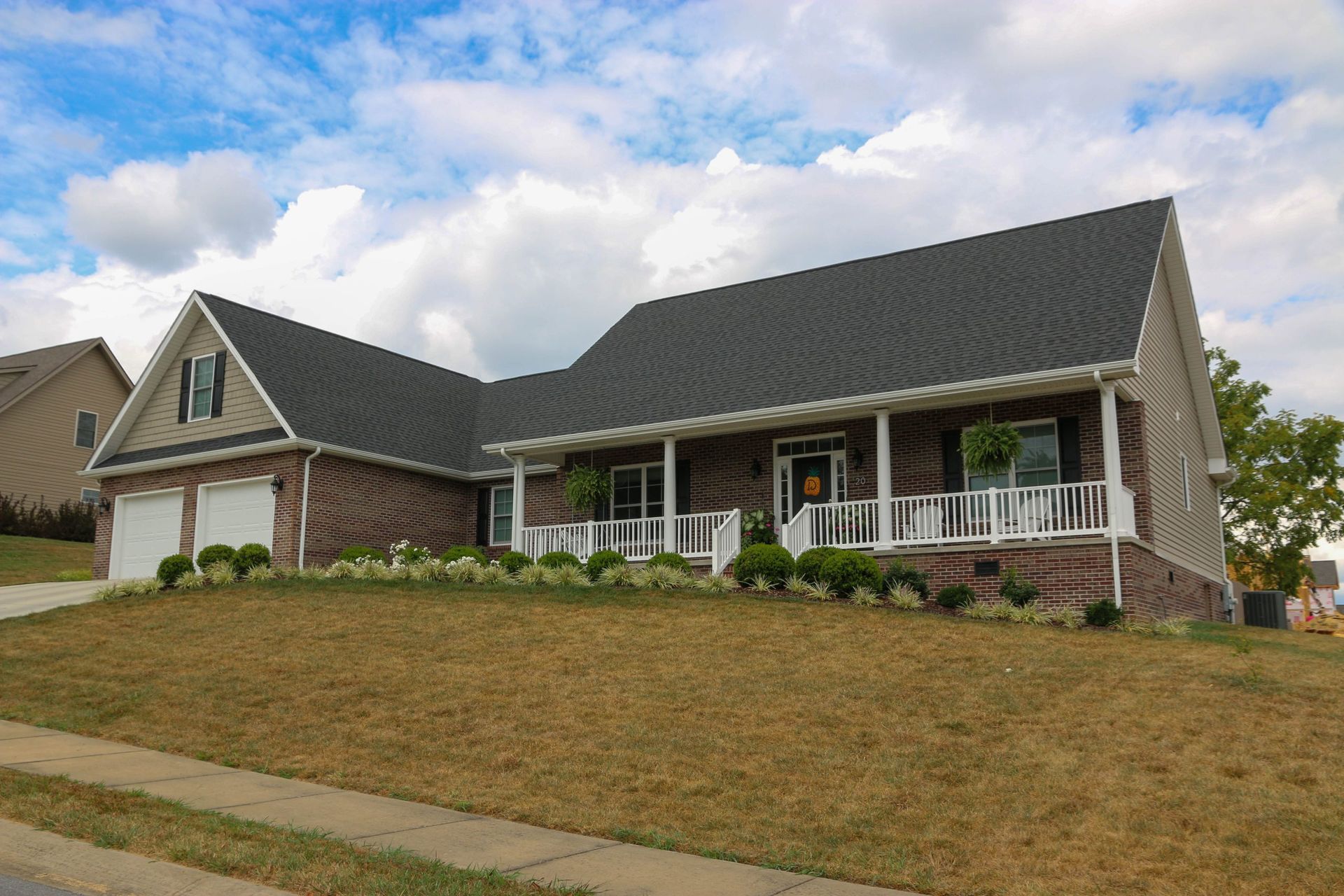 Suburban brick house with a dark roof, white porch columns, and a sloping grassy lawn under cloudy skies