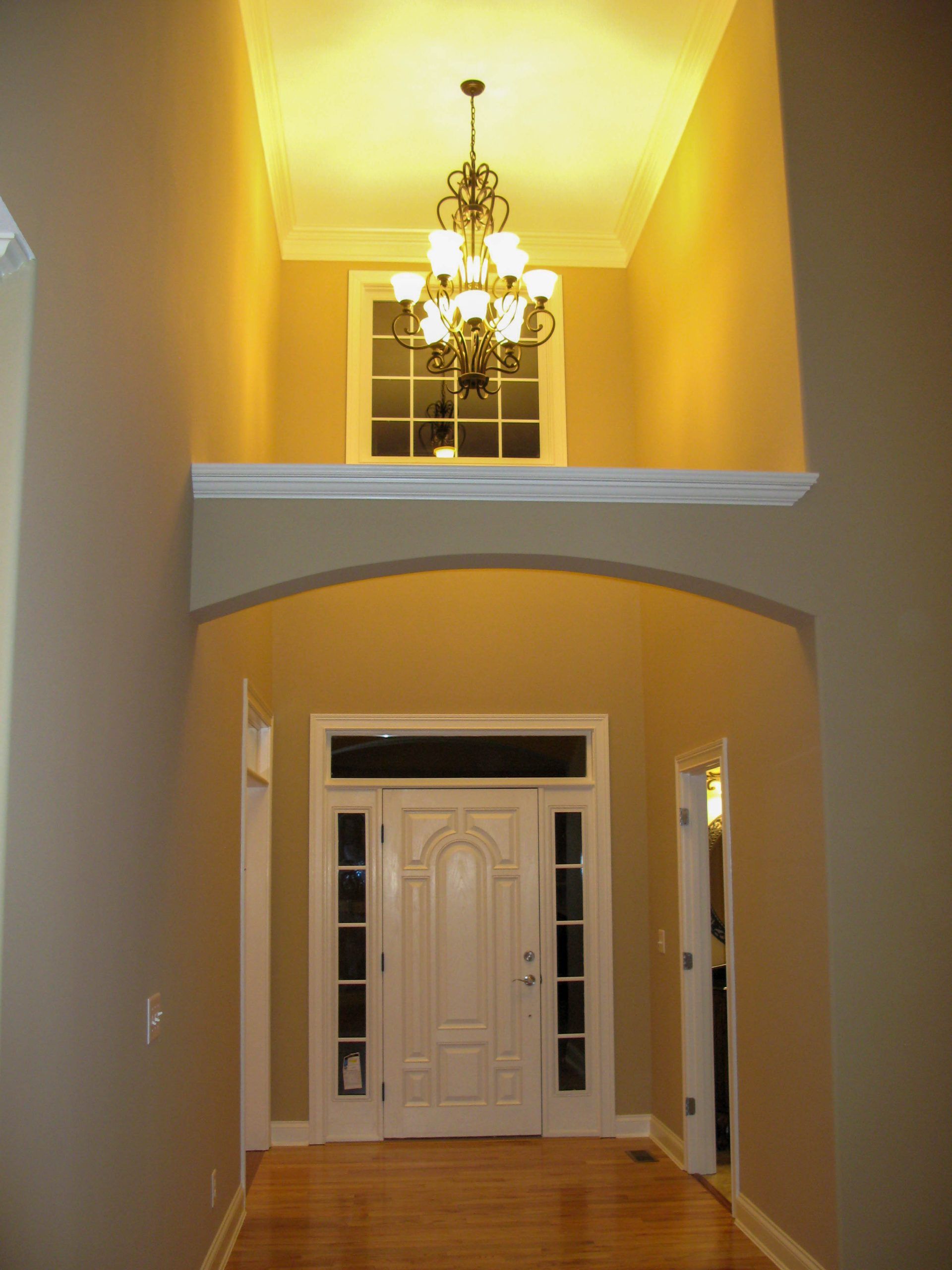 Warm-lit entryway with a white front door, arched hallway, and chandelier above a wood floor.
