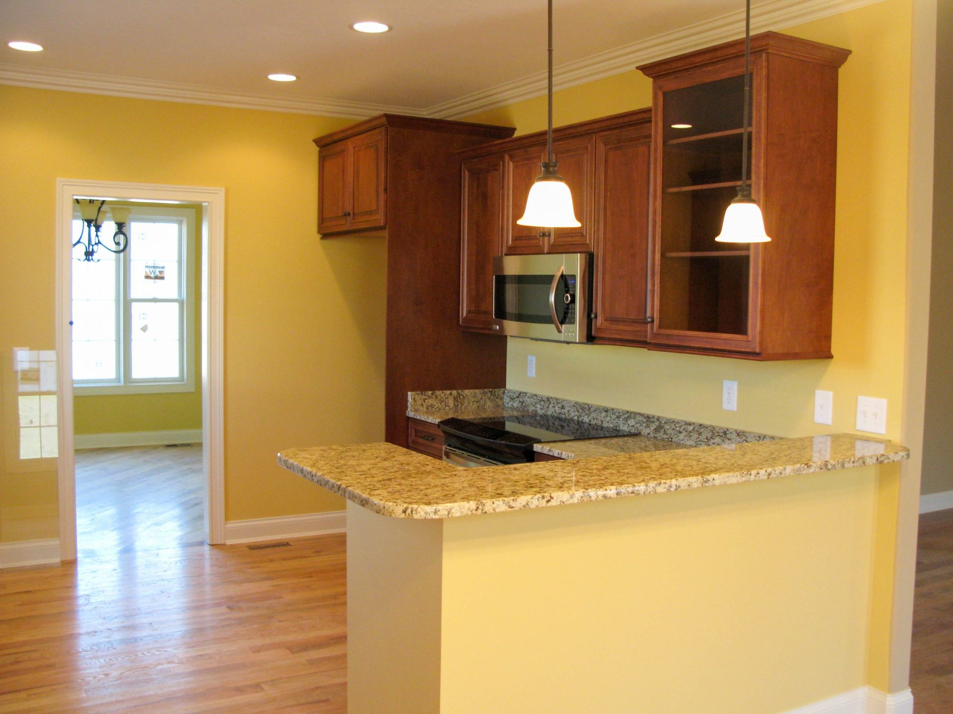 Kitchen with granite island, warm wood cabinets, pendant lights, and a view into a bright adjoining room.