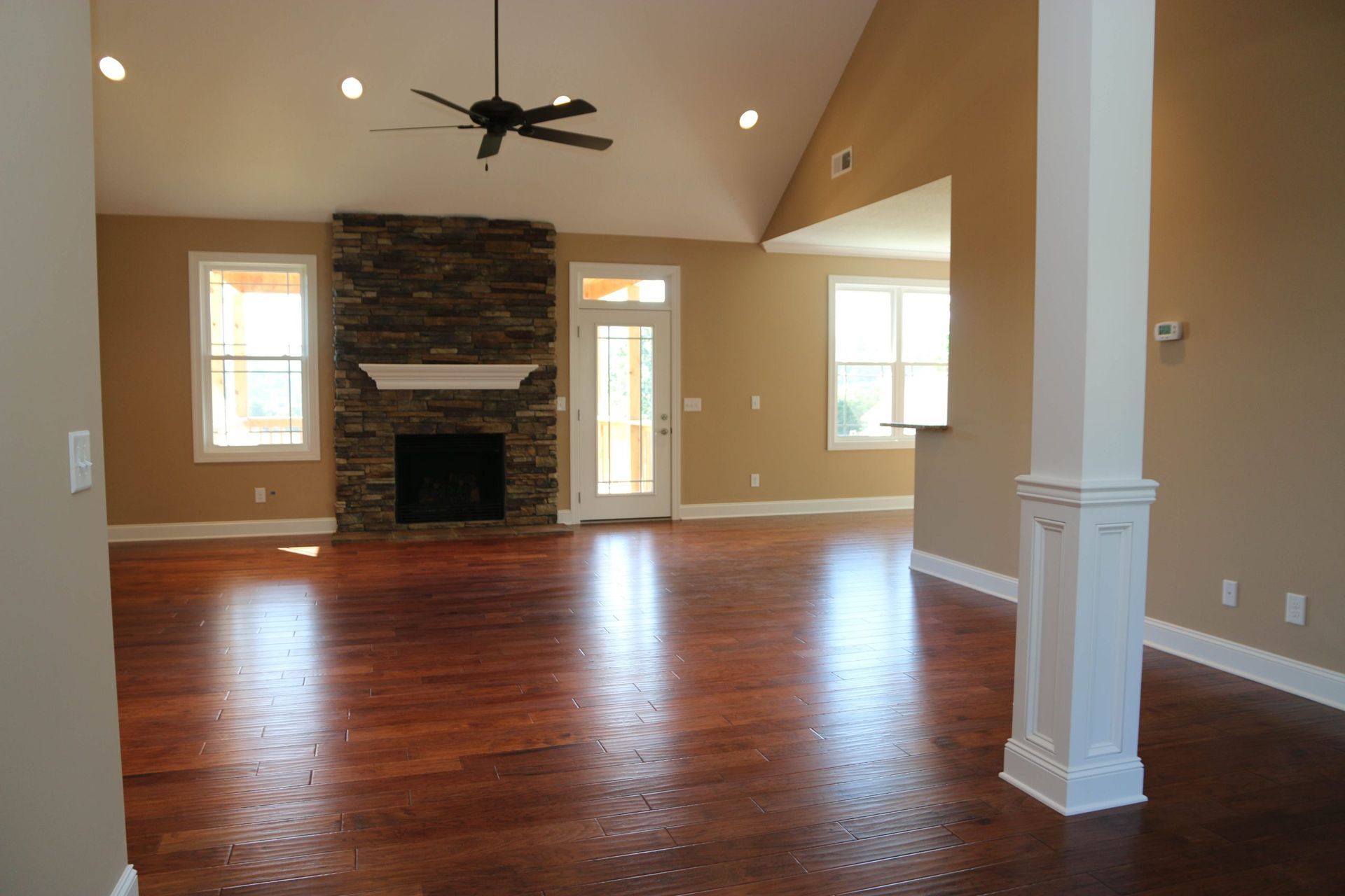 Empty living room with hardwood floors, stone fireplace, beige walls, and a ceiling fan.