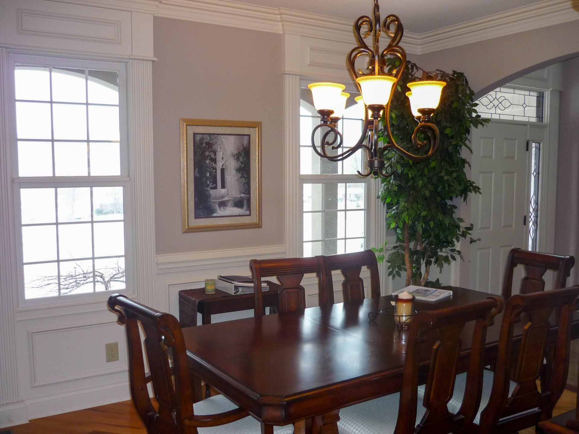 Dining room with dark wood table, chandelier, white wainscoting, large windows, and a potted plant.