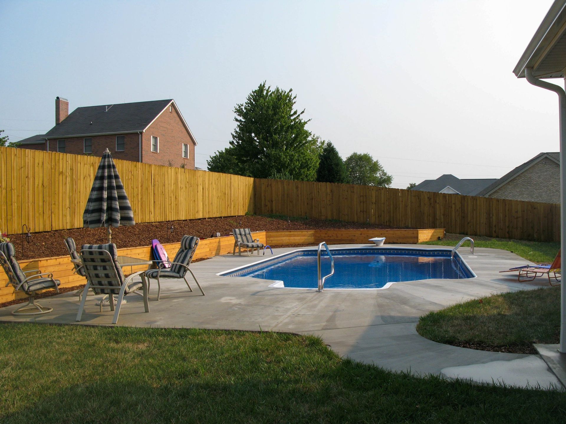 Backyard patio with an in-ground pool, lounge chairs, umbrella, and wooden fence under a cloudy sky