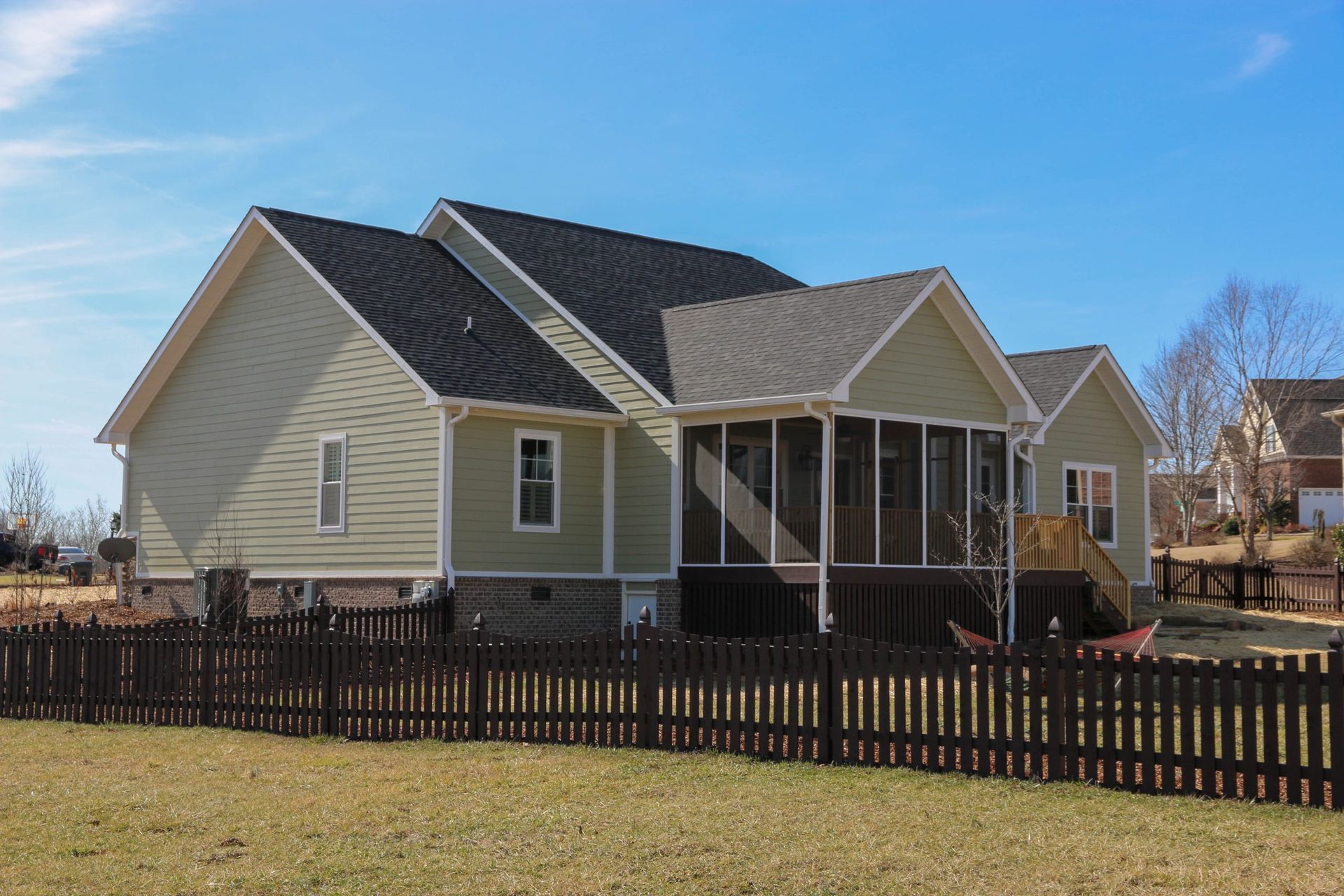 Suburban house with beige siding, dark roof, screened porch, and black picket fence under a blue sky