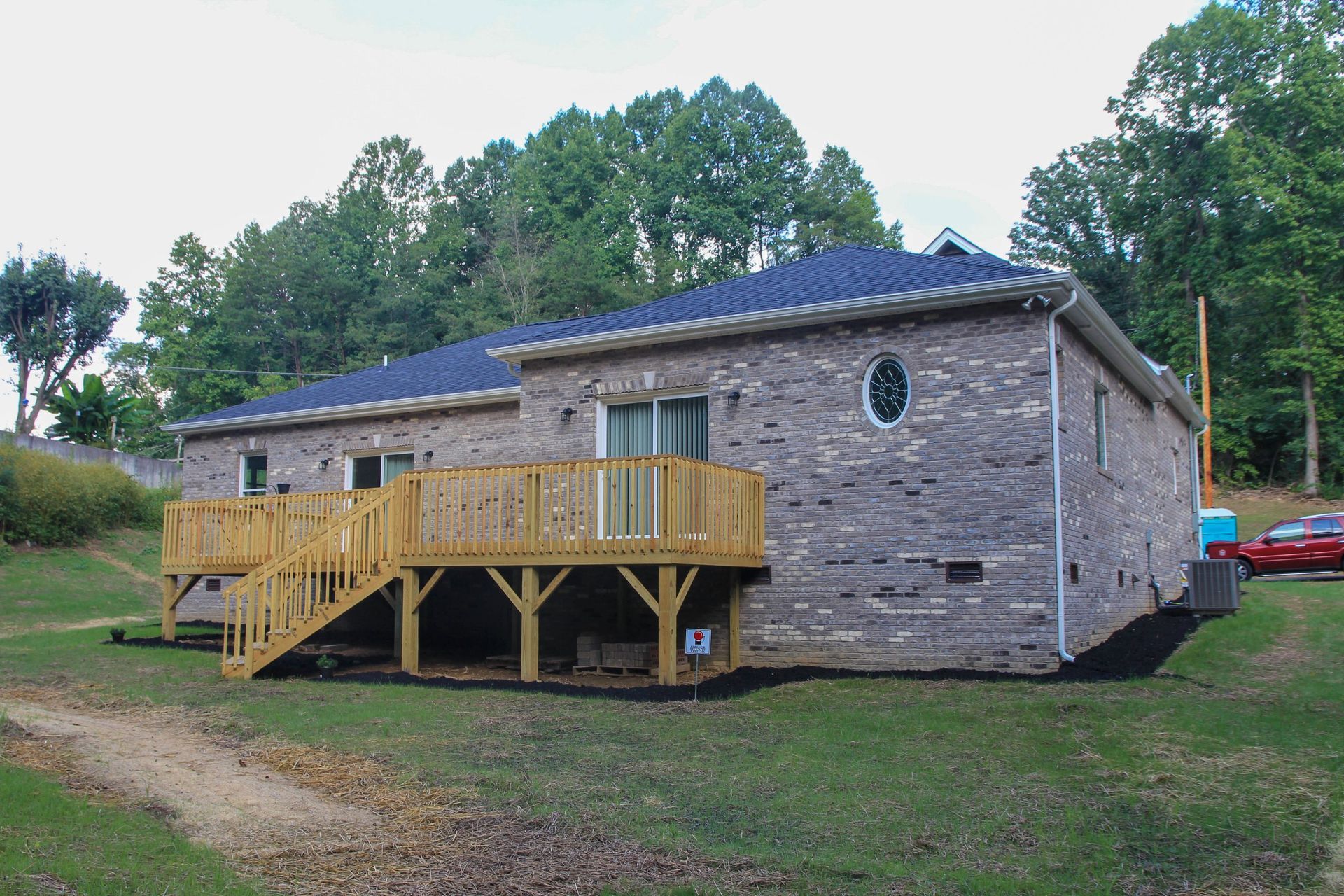 Stone house with a wooden deck and stairs, surrounded by grass and trees